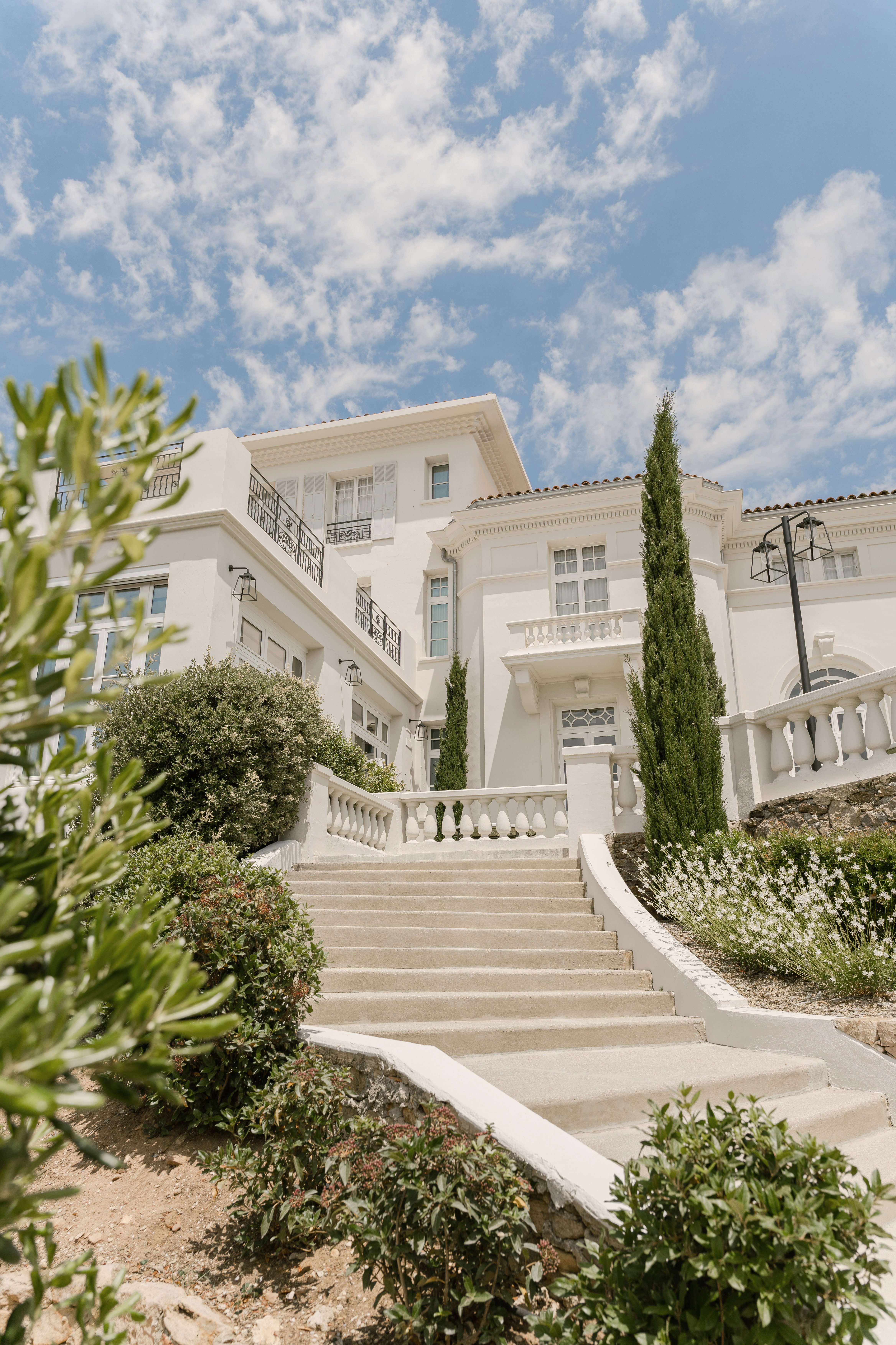 Low-angle view of white multi-storey villa with balustrade stairs, cypress trees, and wrought-iron balconies