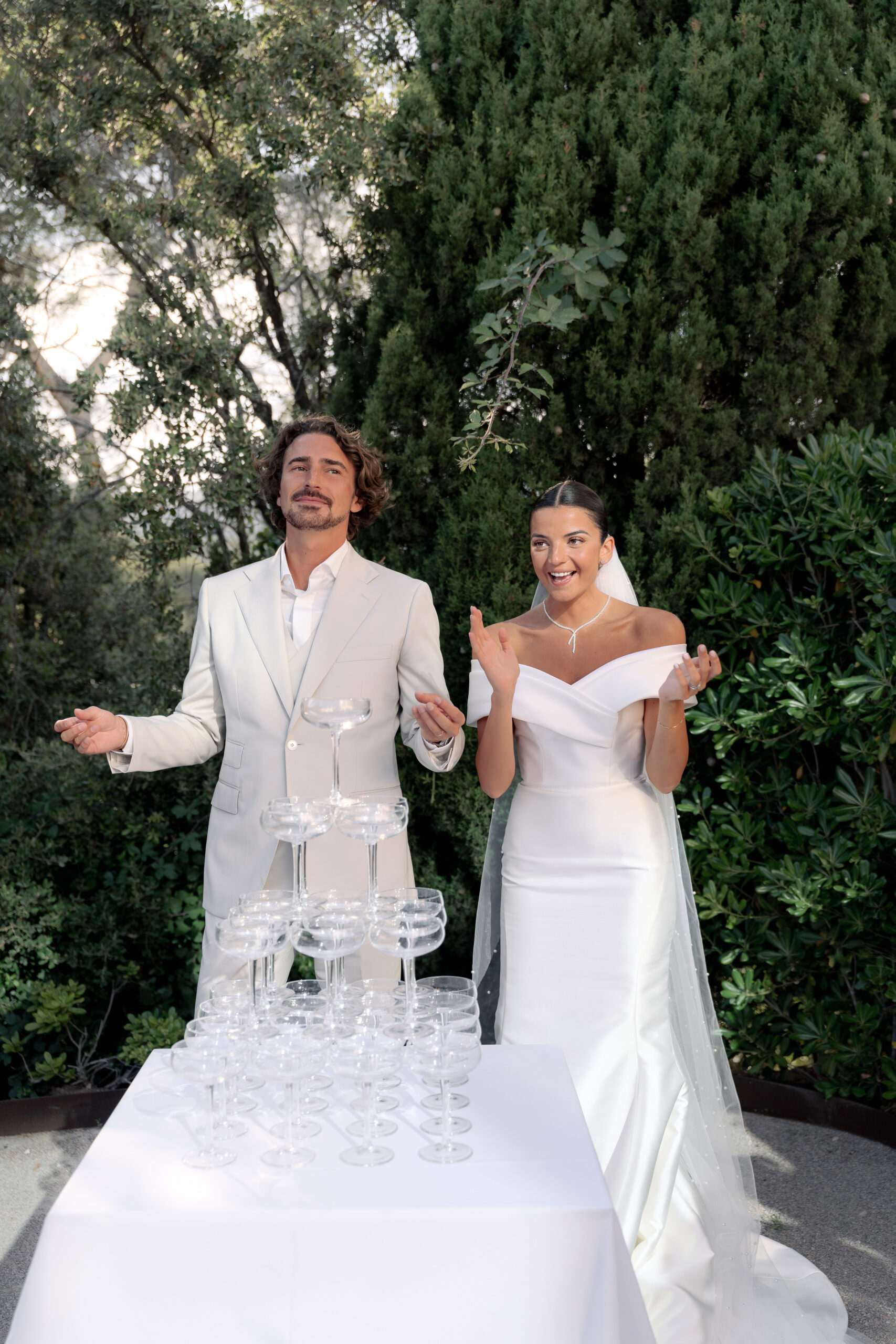 Couple reacts playfully at champagne coupe tower on white-clothed table on garden terrace