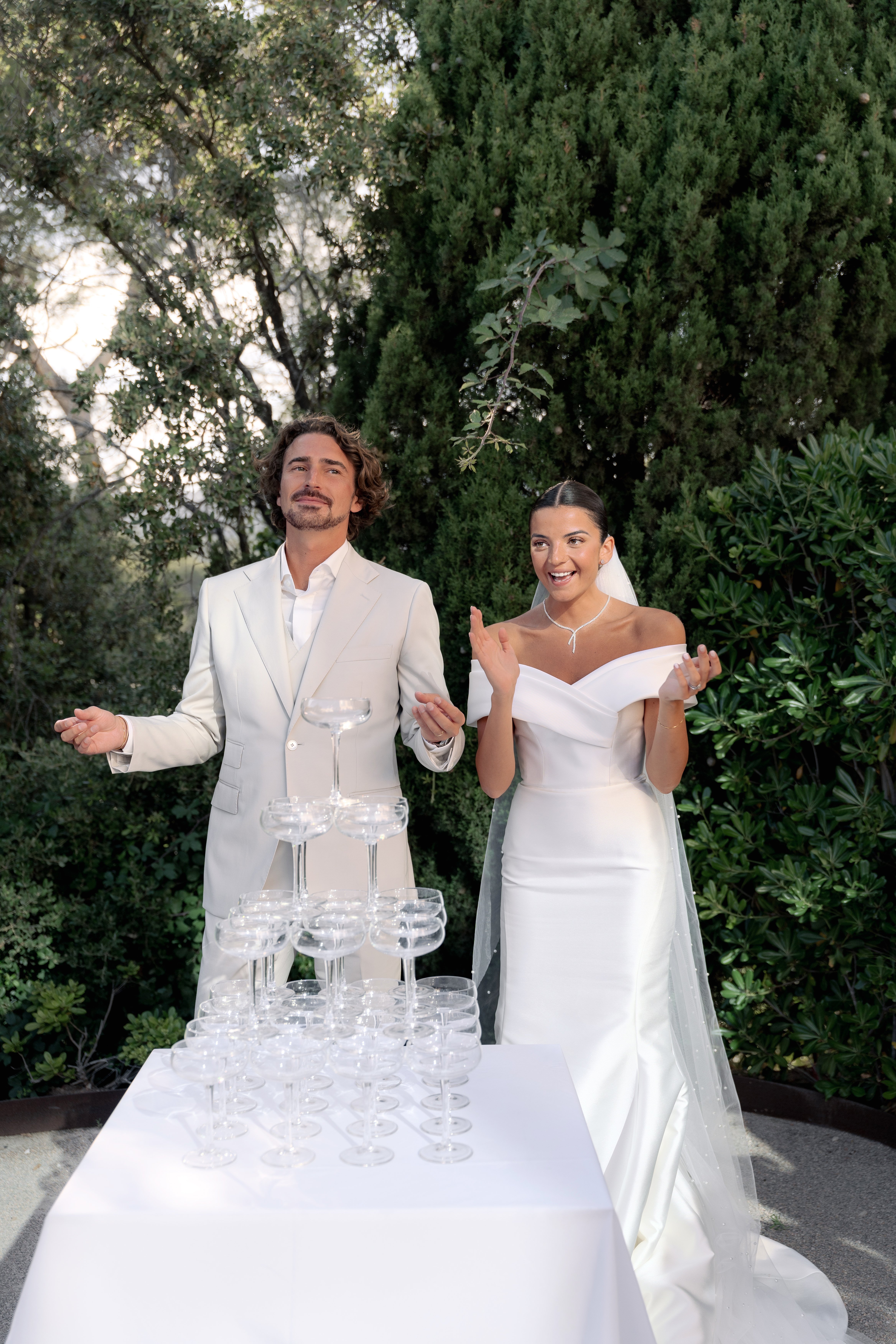The bride and groom stand outdoors behind a white-clothed table set with a champagne tower of coupe glasses, appearing to react with animated expressions just before or after pouring. The groom wears a sand-colored suit with an open-collar white shirt, while the bride wears an off-the-shoulder fitted ivory satin gown with a long veil and a diamond necklace. The setting is an outdoor terrace or garden area with dense cypress and mixed greenery in the background, suggesting a modern, minimalist aesthetic with a clean white-on-white color palette. The shot is a medium portrait-style image capturing both figures at three-quarter length alongside the full champagne tower.