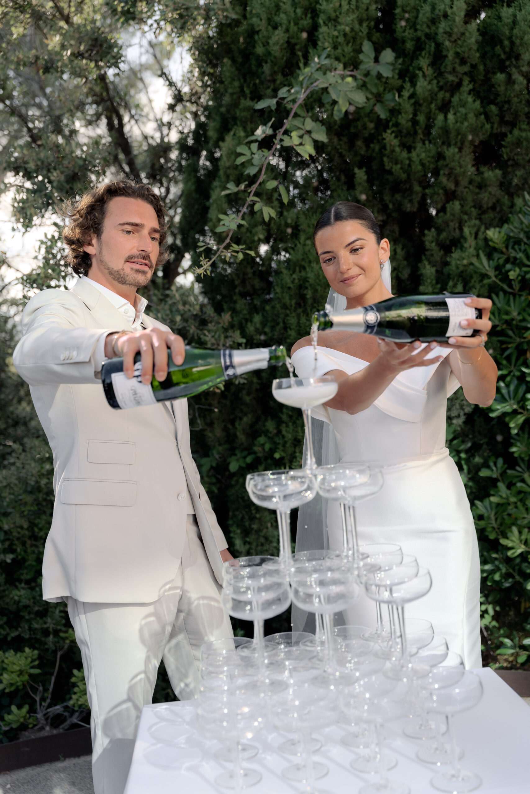 Bride and groom pouring champagne into coupe glass tower outdoors in matching white attire