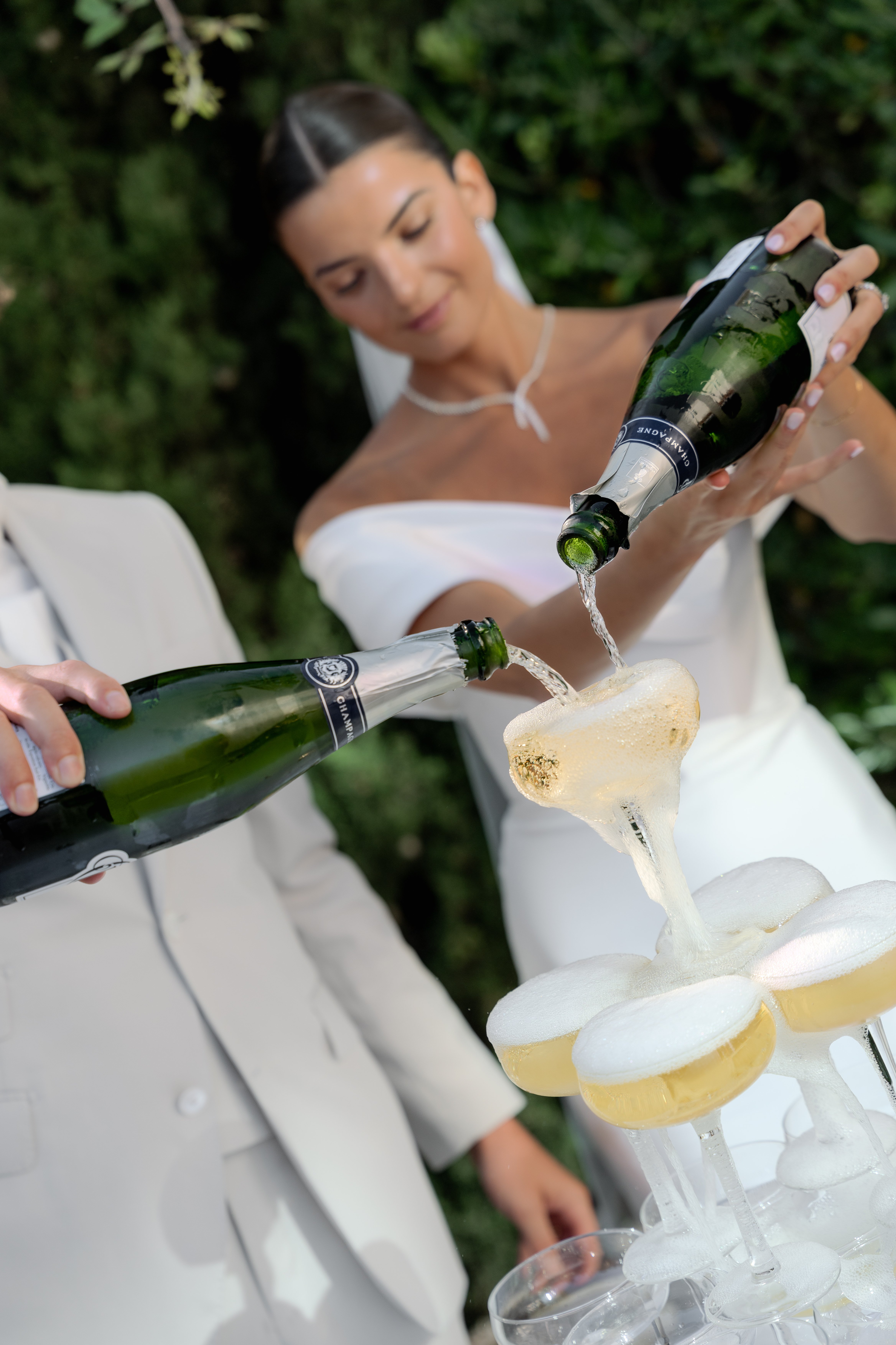 The bride and groom are pouring champagne simultaneously into the top tier of a champagne tower during what appears to be a cocktail hour or reception moment outdoors. The bride wears a white strapless fitted dress with a delicate white necklace and has her dark hair pulled back sleekly; the groom is in a light ivory or off-white suit. Both are holding dark green champagne bottles labeled 'Champagne,' and the liquid is visibly overflowing and cascading down into the coupe glasses below. The tower consists of multiple tiers of wide champagne coupe glasses filled with golden, frothy champagne. The shot is a close-up portrait-style image with the action in sharp focus in the foreground and the bride softly blurred in the background.