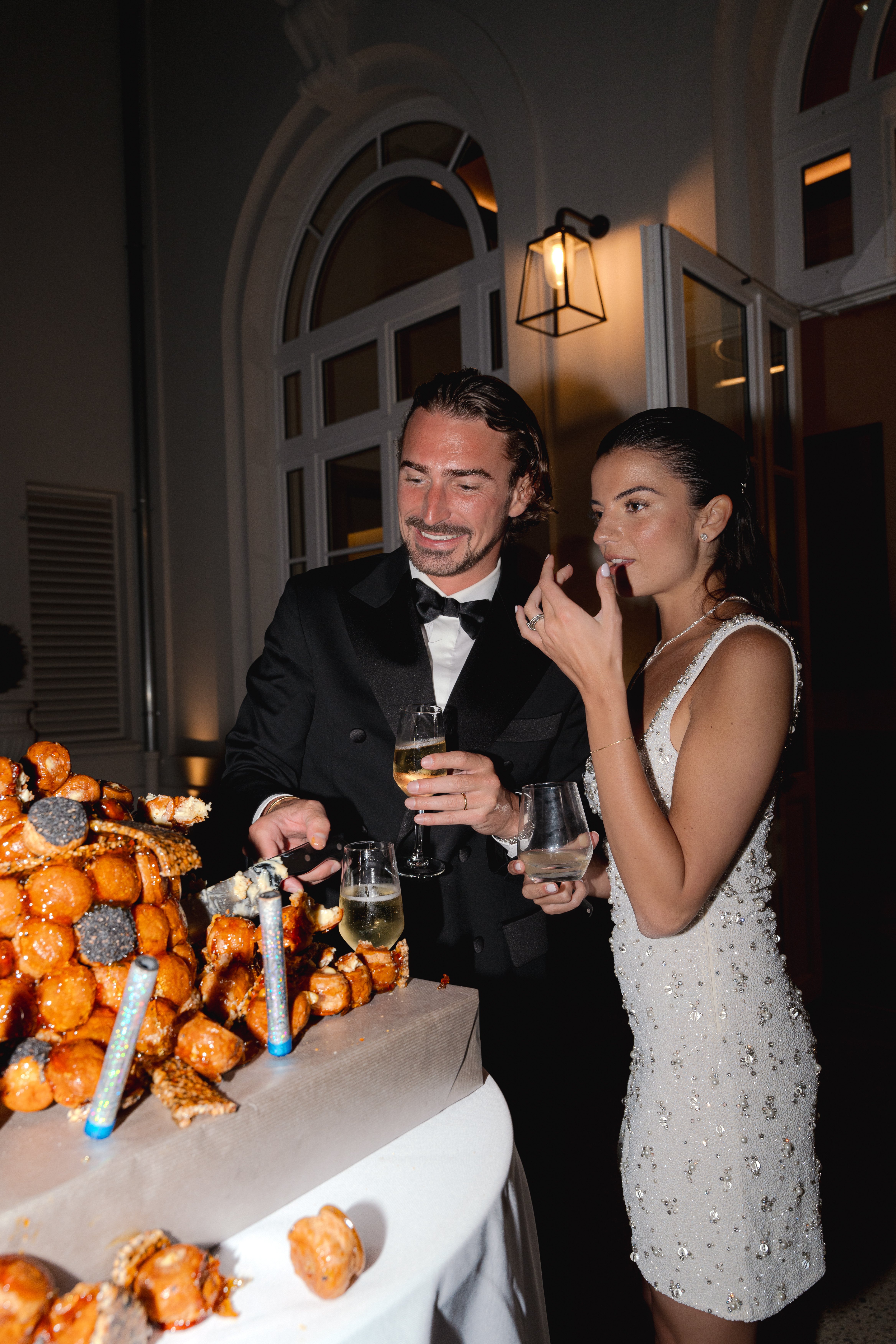 The couple is cutting into a croquembouche — a traditional French choux pastry tower — during what appears to be an evening reception. The groom wears a black double-breasted tuxedo with a bow tie, and the bride has changed into a short white mini dress covered in silver beading and crystal embellishments, suggesting this is a reception outfit change. Both are holding champagne glasses, and the bride is tasting a profiterole. The setting is an outdoor or semi-covered terrace area at night, lit by a wall-mounted black lantern and warm ambient uplighting, with arched white-framed windows and doors visible in the background. The croquembouche is decorated with dark chocolate accents and sparkler-style candles. The shot is a medium portrait-style image capturing a candid, relaxed moment between the two.
