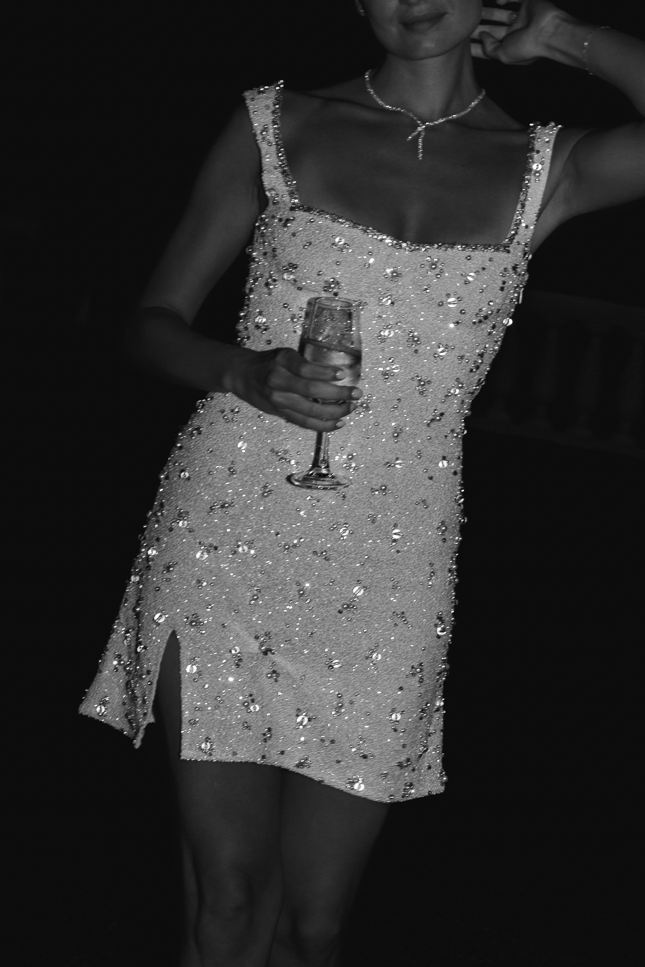 A black-and-white close-up portrait of a woman, likely a guest or bride in a reception outfit change, holding a champagne flute at what appears to be an evening wedding reception. The image is cropped from chin to mid-thigh, showing a short, square-neck mini dress heavily embellished with sequins, beading, and scattered rhinestones, with a small front slit at the hem. She wears a delicate chain necklace and her hand rests near her chin. The background is very dark, suggesting a dimly lit indoor evening setting. The high-contrast black-and-white tones emphasize the reflective texture of the beaded dress against the near-black background.