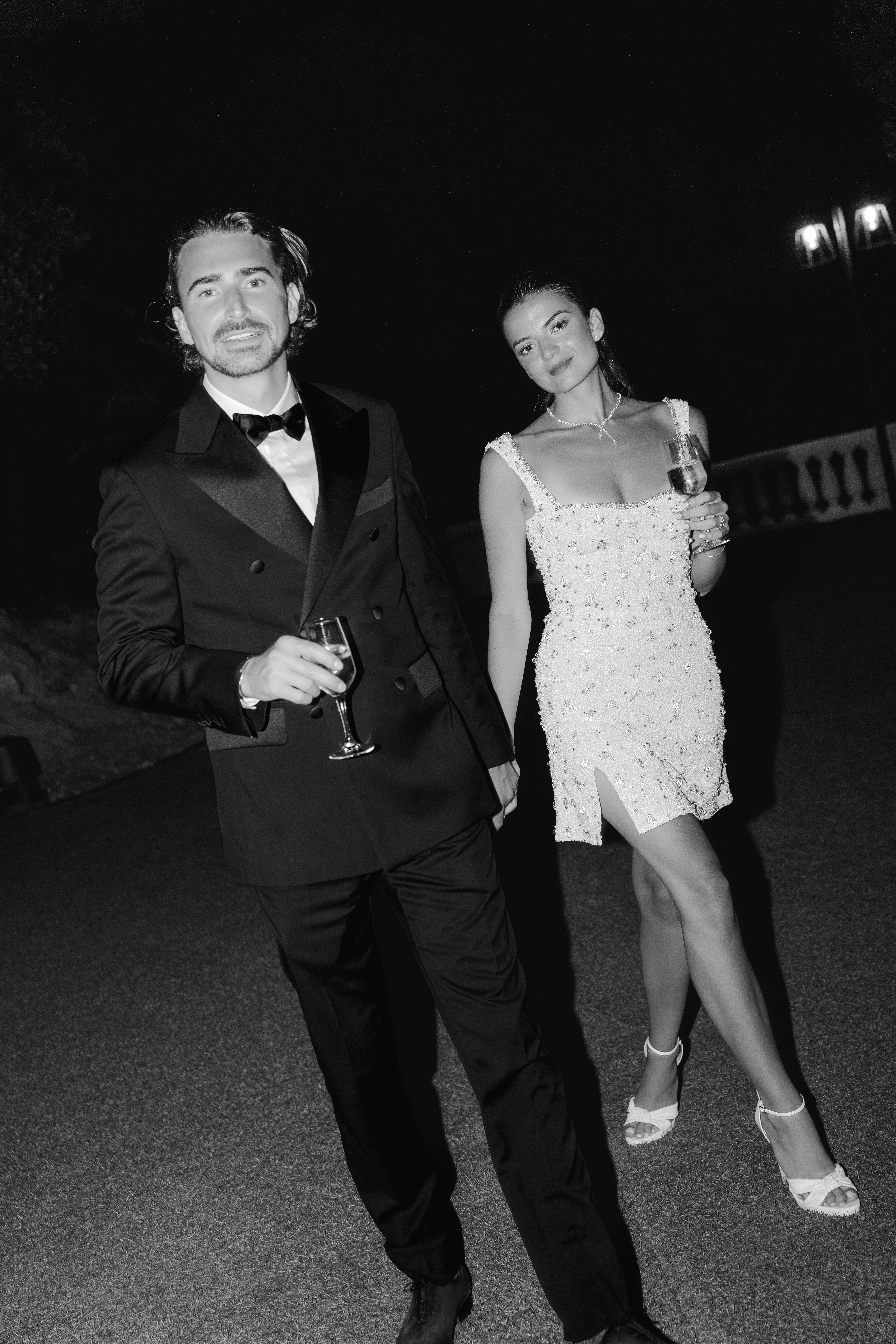 Couple holding champagne flutes at night, groom in tuxedo and woman in embellished mini dress in B&W