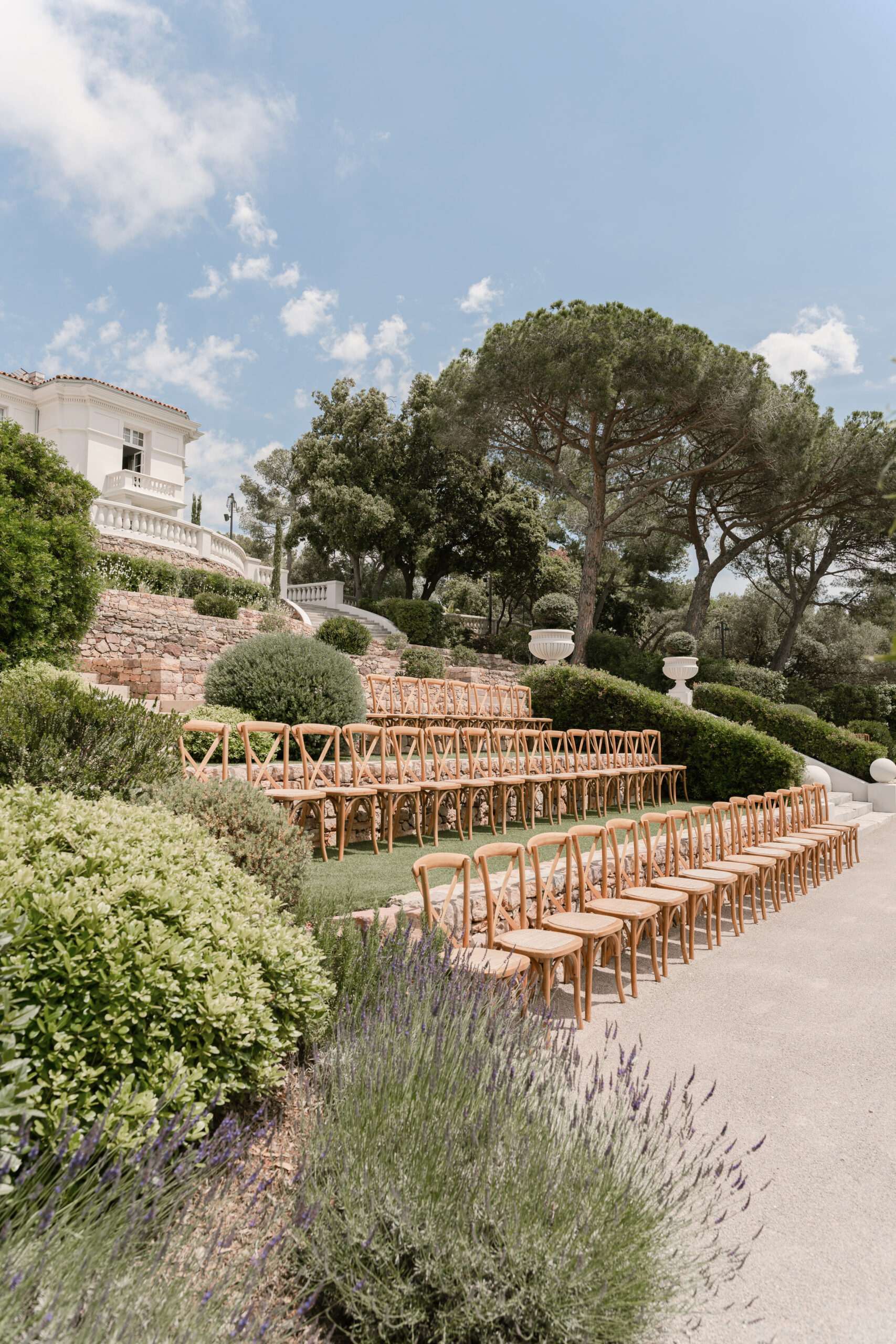 An outdoor wedding ceremony setup photographed before guests arrive, shot from a wide angle showing the full seating arrangement. Rows of natural wood cross-back chairs are arranged in two sections with a central aisle on a gravel terrace, accommodating approximately 60–80 guests. The setting is a formal terraced garden at a white classical villa, with a stone retaining wall, white balustrades, and white decorative stone urns visible on the upper level. Lavender plants in bloom line the foreground of the seating area, and the overall decor aesthetic is classic Provençal with no added floral or fabric chair decorations. Potential venue feature image.
