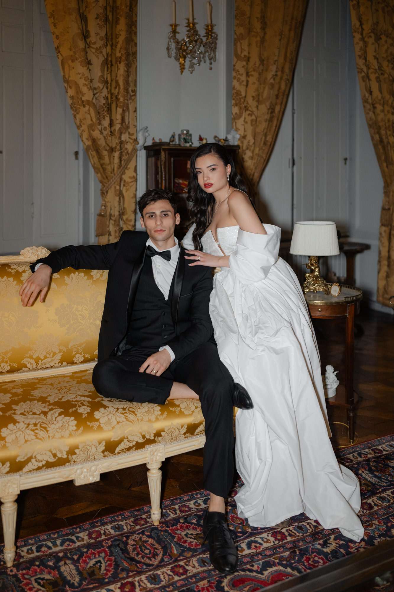 Bride and groom seated on gold damask sofa in classical interior with chandelier and ornate gold furnishings