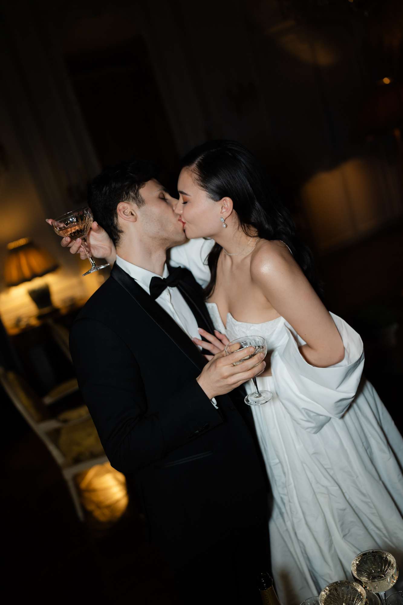 Bride and groom kissing while holding champagne flutes during nighttime reception with warm amber lighting