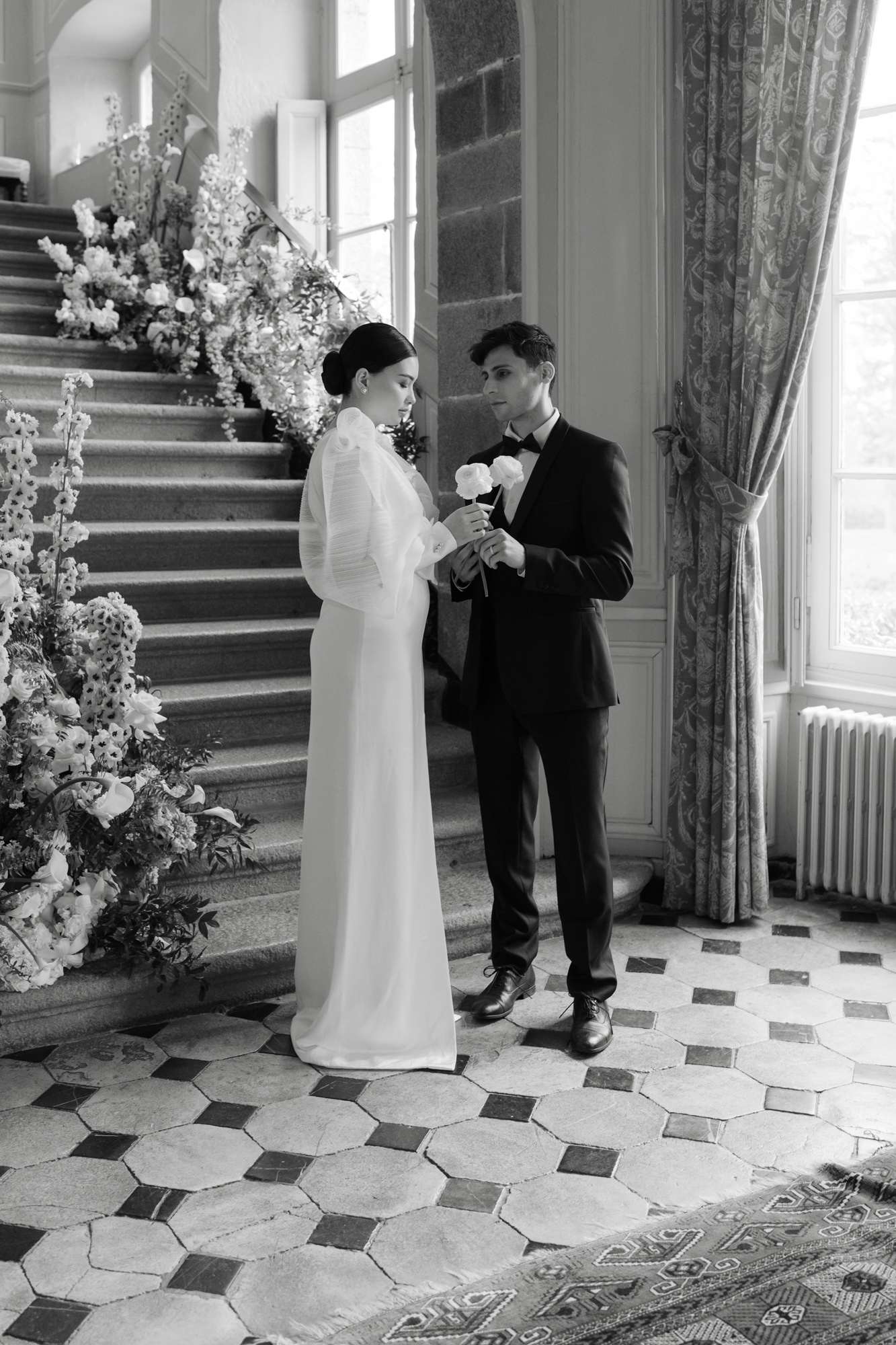 Black-and-white portrait of couple on grand staircase with cascading white floral installation and marble floor