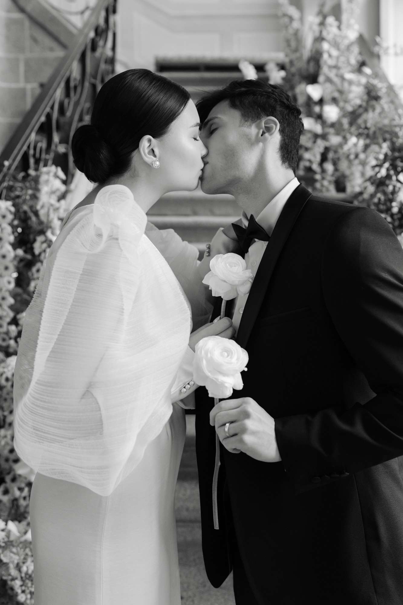 Black-and-white bride and groom kissing in corridor flanked by tall white floral installations holding roses