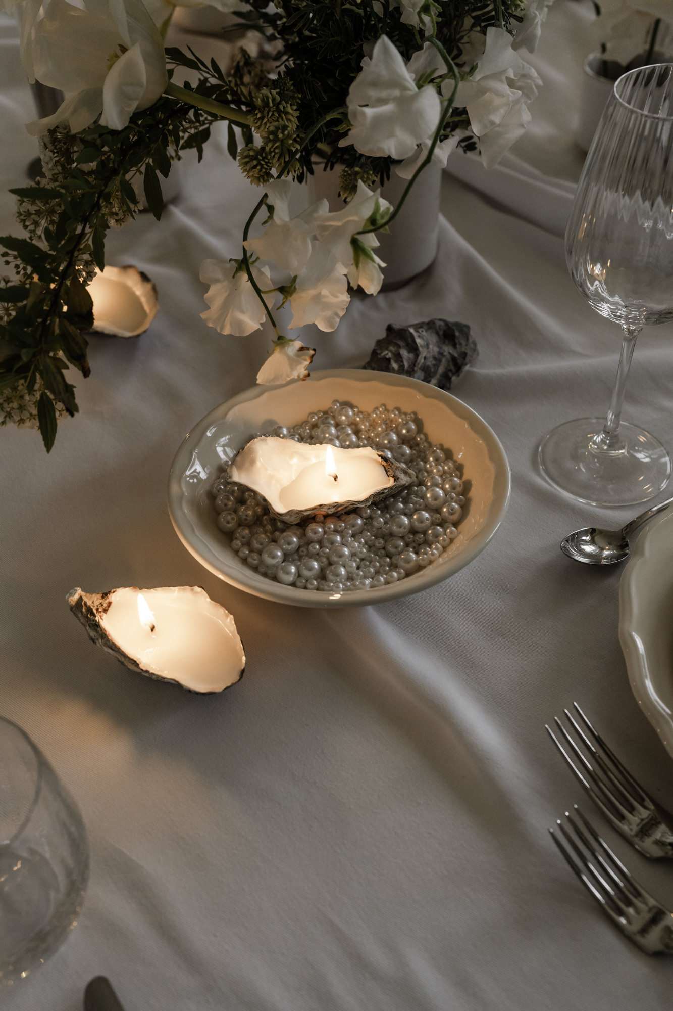 Reception place setting with oyster shell candle, pearl-bead glass charger, crystal glassware and white sweet pea floral arrangement.