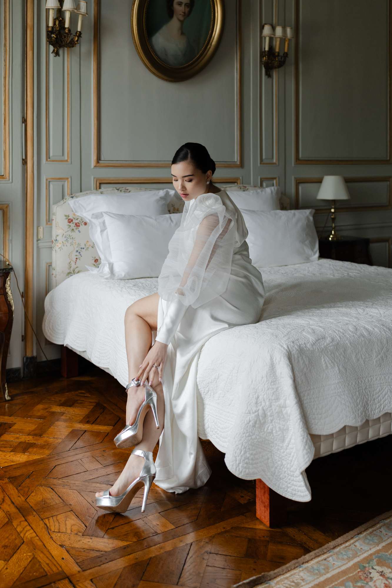 Bride adjusting silver heels on bed in classical French bedroom with gilt frames and sage walls