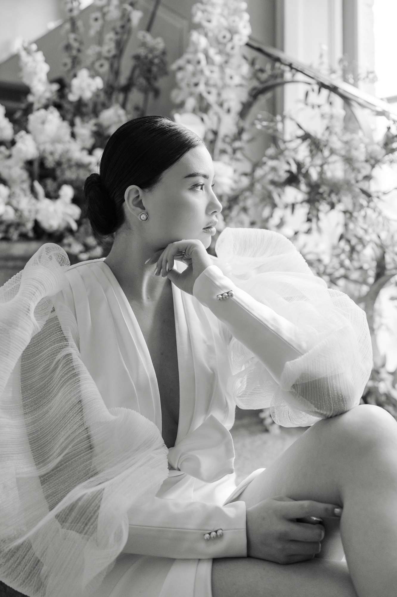 Black and white portrait of bride in structured white blazer seated in profile before indoor white floral installation