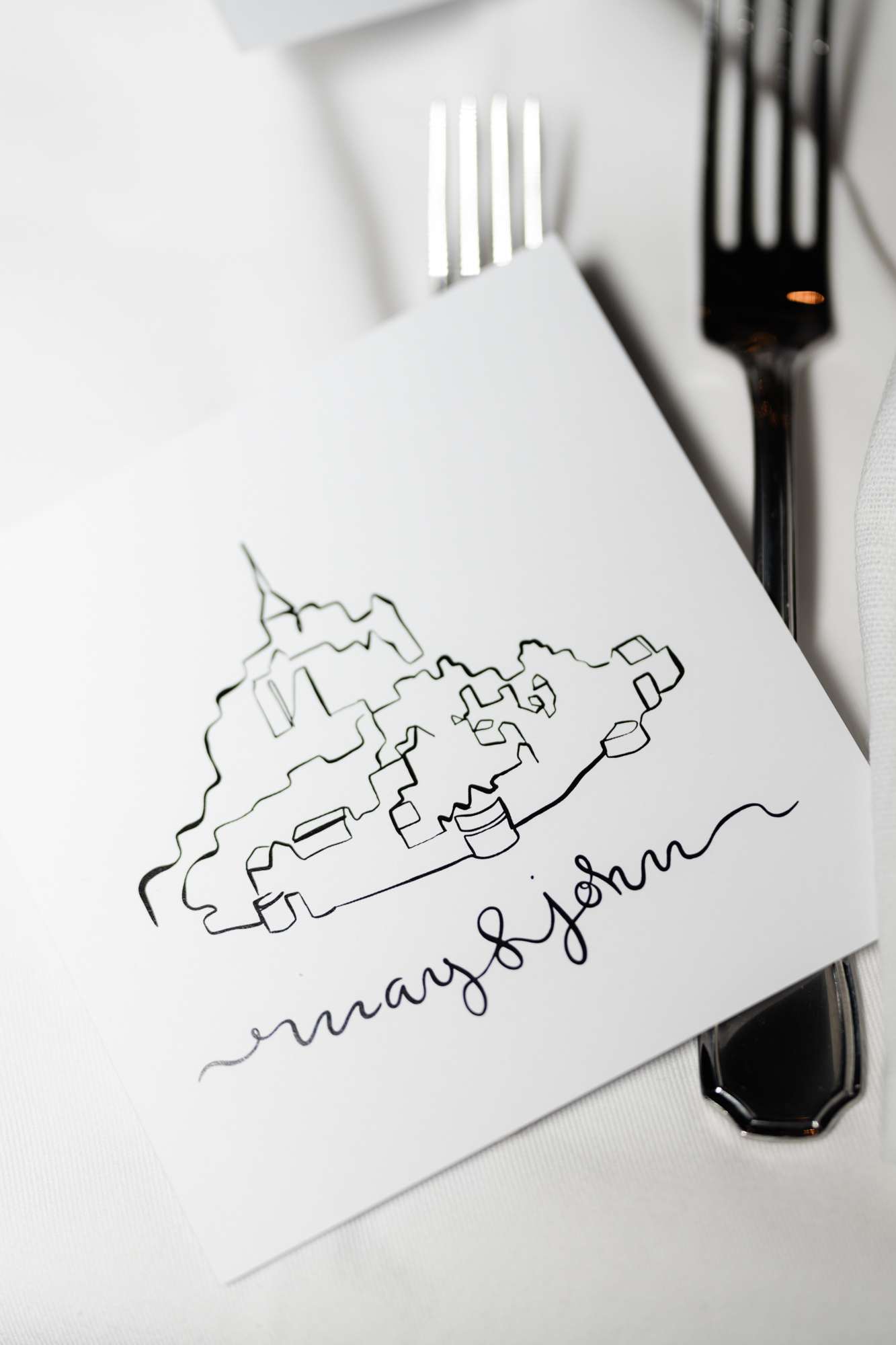 Hand-drawn place card with architectural illustration on white napkin beside fork and plate at reception table