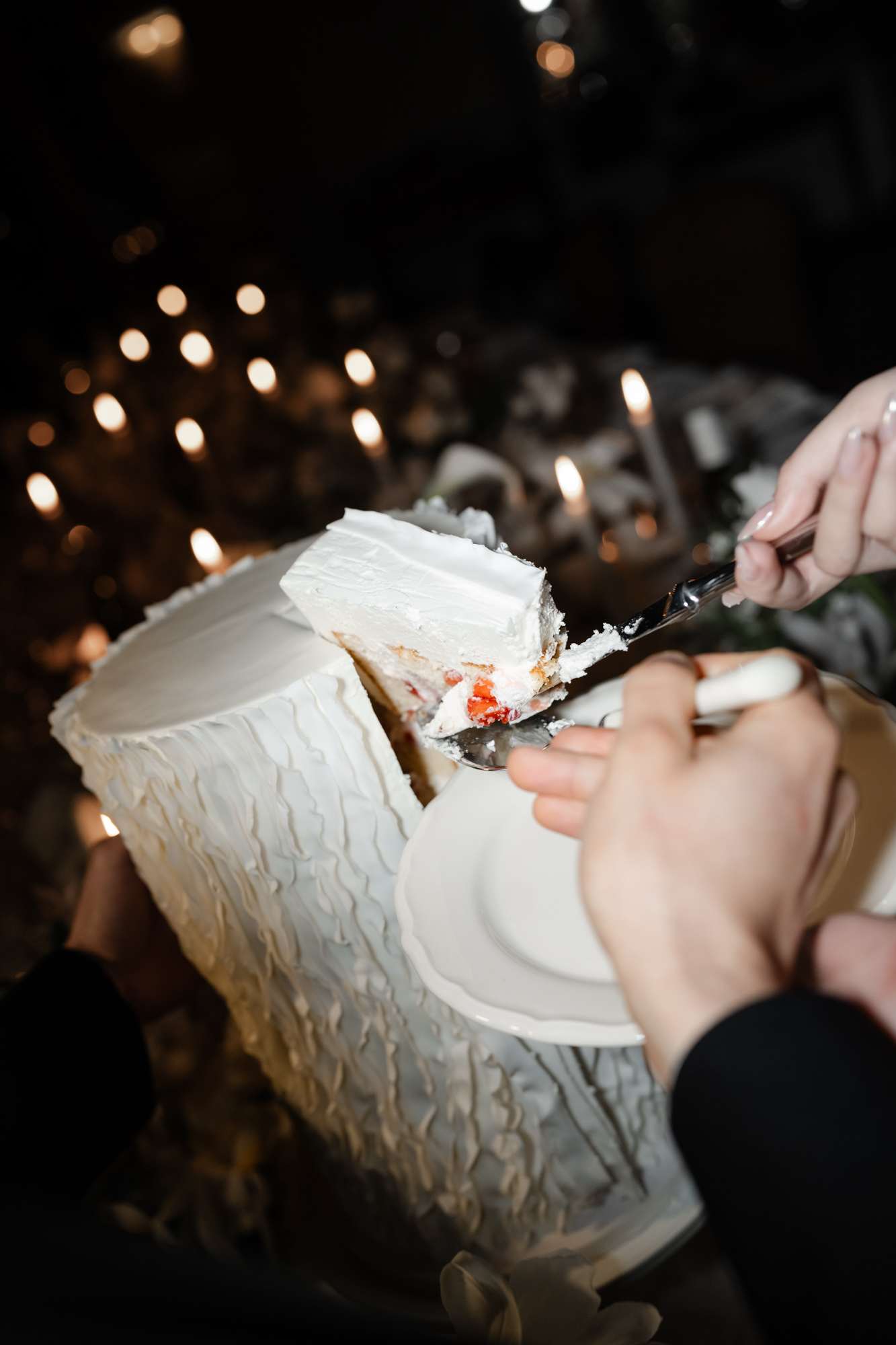 Hands lifting cake slice with ivory buttercream exterior and red fruit filling in candlelit reception