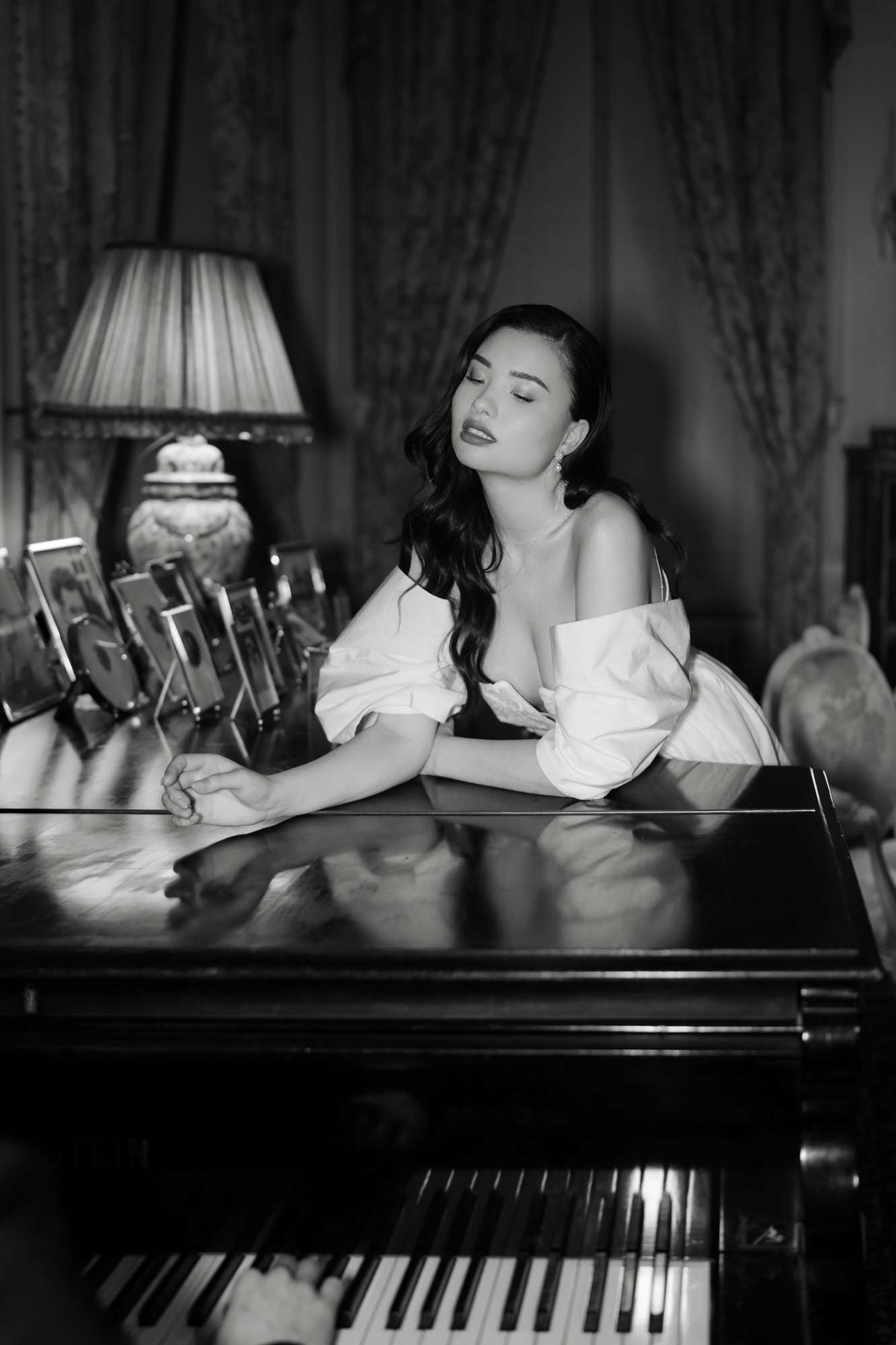 Bride in off-shoulder white gown seated at grand piano in classical interior with dark curtains, black and white
