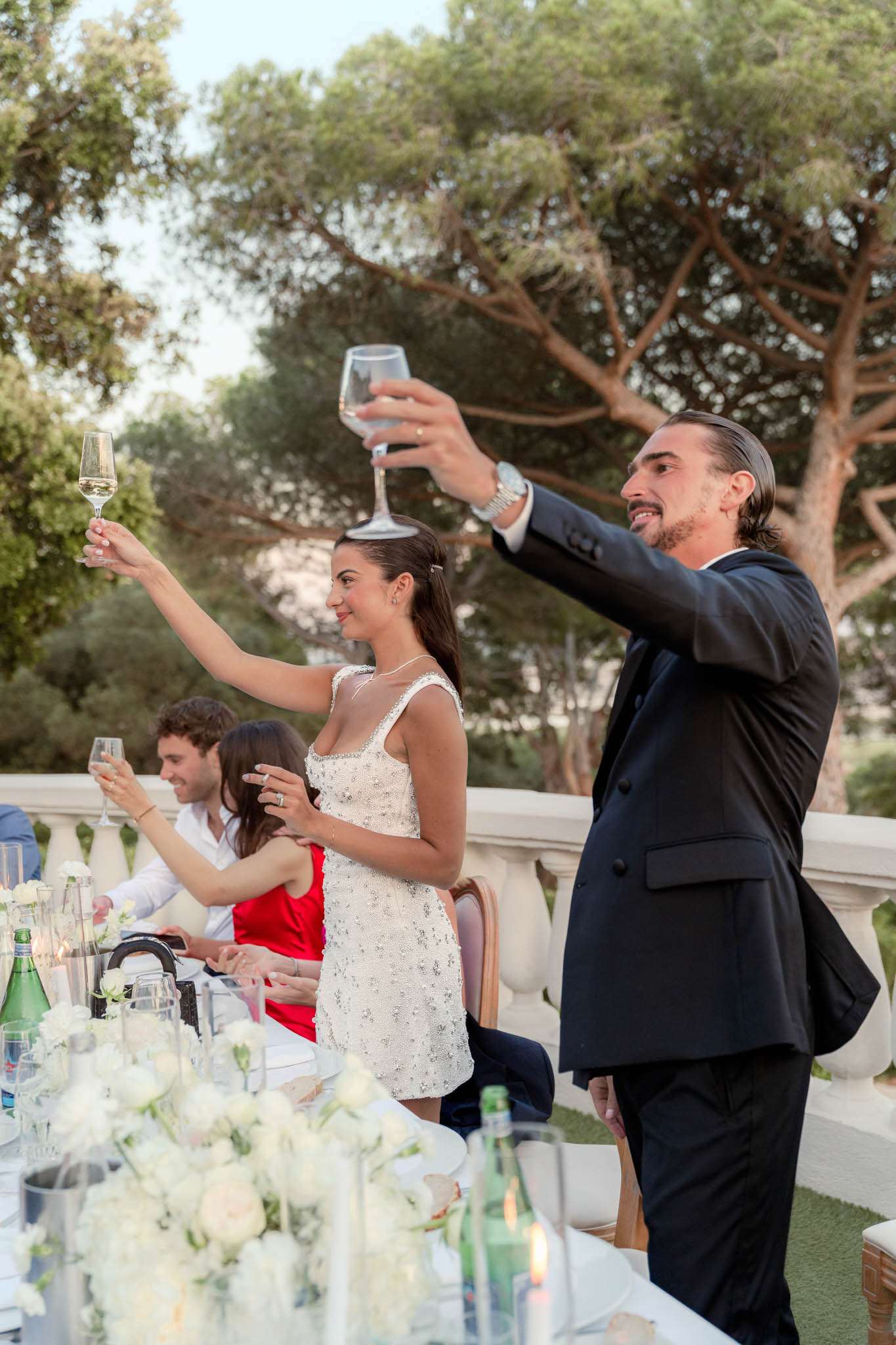 Bride and groom raising champagne glasses during outdoor terrace reception toast with seated guests