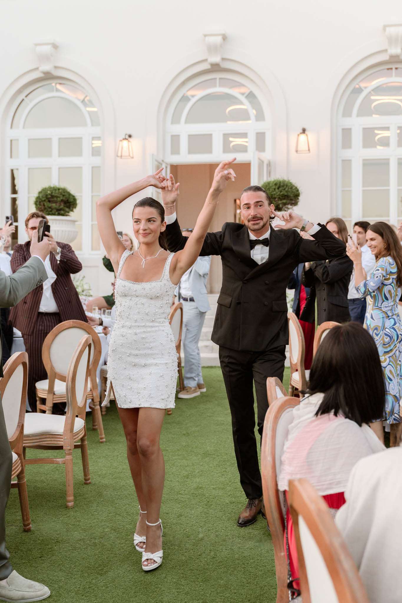 Bride in white beaded mini dress and groom in black tuxedo making grand entrance to outdoor reception as guests cheer