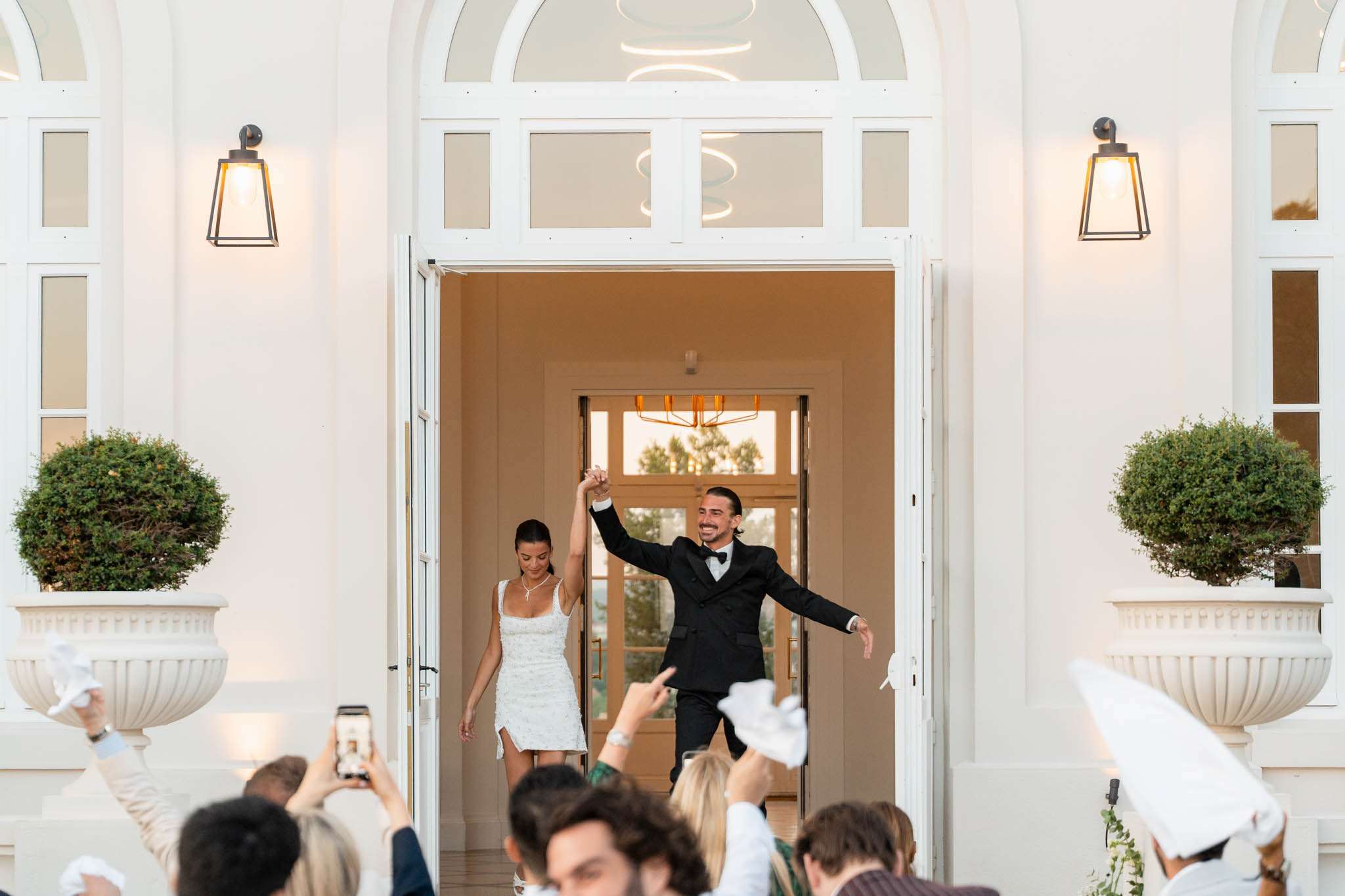 The bride and groom are making their grand entrance through the main double doors of a white neoclassical villa or château, with guests in the foreground cheering, waving white napkins or handkerchiefs, and photographing with phones. The groom, in a black tuxedo with a bow tie, raises the couple's joined hands triumphantly while extending his other arm outward; the bride wears a short white fitted dress with a square neckline and subtle embellishment, accessorized with a delicate necklace. The venue façade is white with arched transom windows, black wall-mounted lanterns, and large white stone urns planted with clipped round topiaries, giving the setting a classic, formal aesthetic. The shot is taken from a medium distance at guest level, framing the couple centrally within the doorway with the warmly lit interior visible behind them. Potential venue feature image.