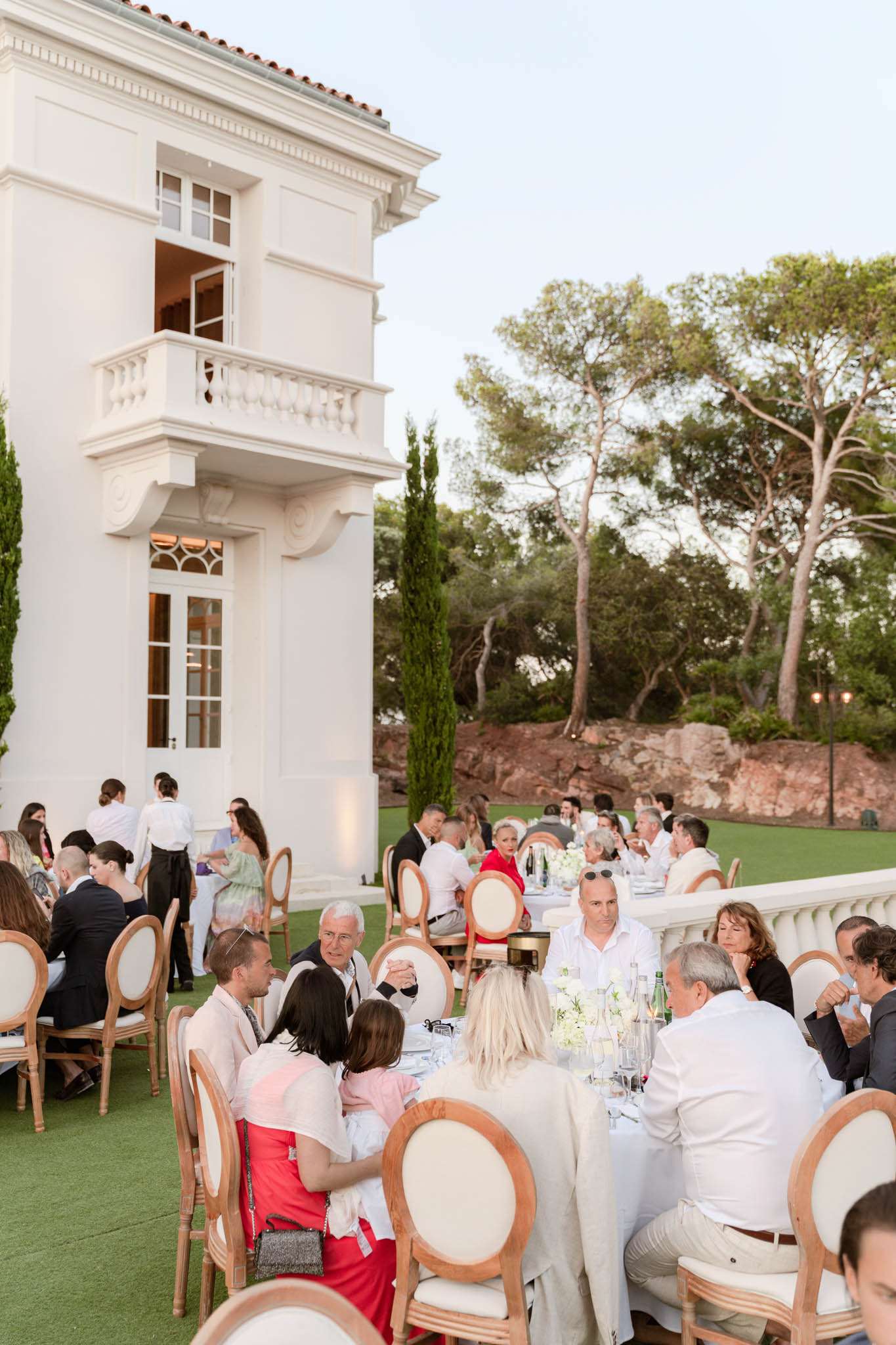 An outdoor wedding reception dinner taking place on a terrace lawn beside a white multi-storey villa with ornate balconies and classical architectural detailing. Approximately 30–40 guests are seated at long rectangular tables dressed in white linen, with centerpieces of white florals and glassware including green-labeled water bottles. Seating consists of Louis XVI-style chairs with natural wood frames and cream upholstery. Guests are dressed in smart-casual to formal attire, with notable pops of red and pastel among the outfits. The table settings and décor follow a clean white palette with minimal color accents. The shot is taken from a slightly elevated angle, capturing both the foreground table and additional tables extending toward the background of the garden. Potential venue feature image.