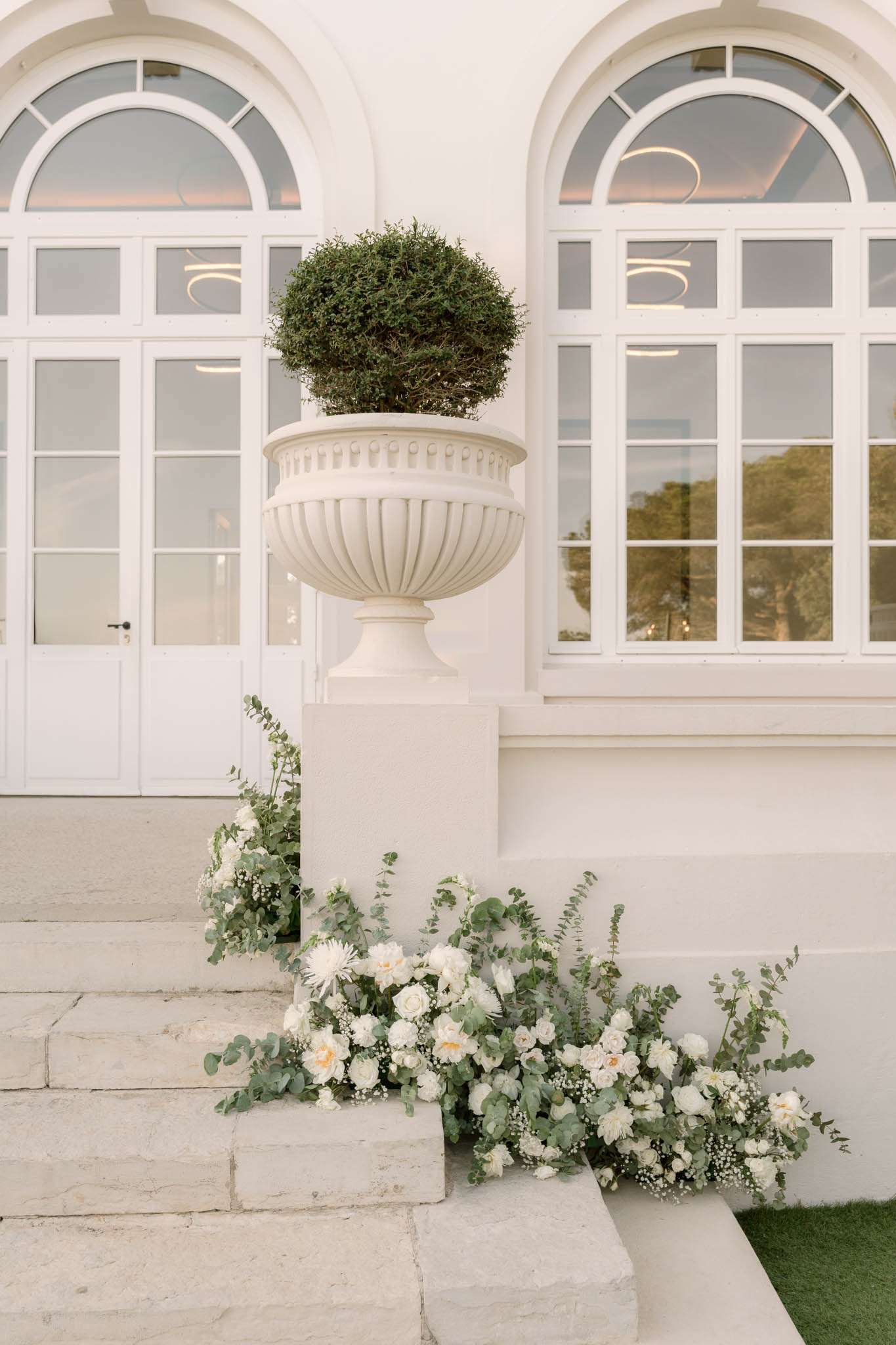 White floral arrangement of roses, dahlias, and eucalyptus at base of stone urn on venue entrance steps