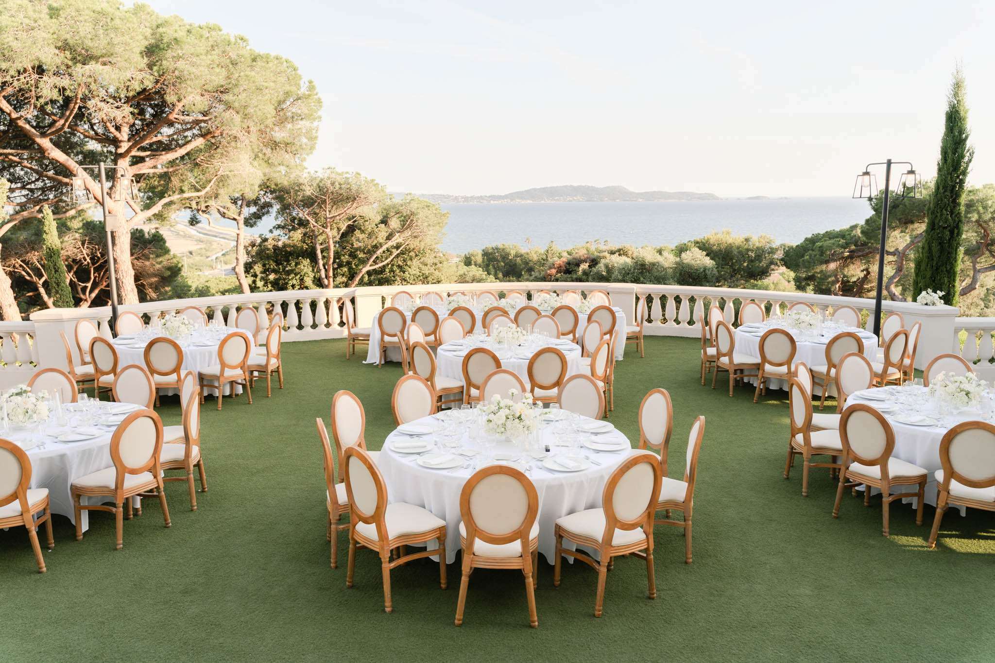 Elevated view of outdoor terrace reception with round white-clothed tables and Louis-style chairs overlooking coastal sea