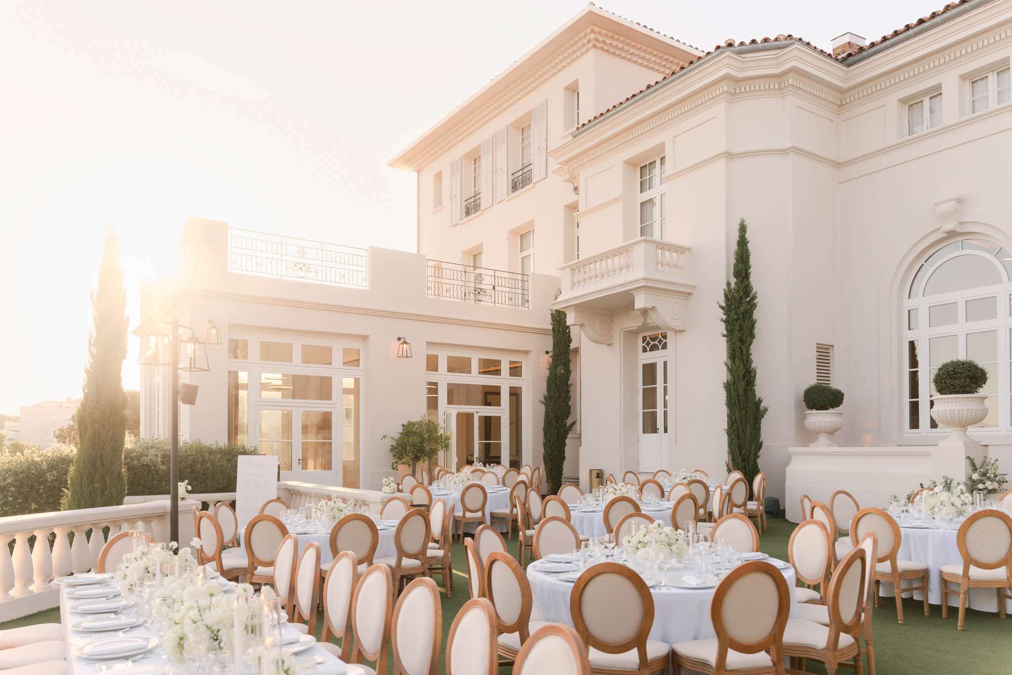 An outdoor wedding reception setup on the terrace of a large white Italianate-style villa, photographed at golden hour with warm backlighting creating a soft, glowing atmosphere. Multiple round dining tables are dressed in pale blue-white linen and arranged across a manicured lawn terrace, each set with white floral centerpieces featuring what appear to be white roses and white blooms with greenery, alongside candles and full glassware and place settings. The chairs are gold-framed Louis XVI-style medallion chairs with cream upholstered seats and backs. The venue building features white render, terracotta roof tiles, wrought-iron Juliet balconies, arched windows, white balustrade railings, tall cypress trees, and large white stone urns with topiary. Wide establishing shot capturing both the full reception layout and the villa architecture. Potential venue feature image.