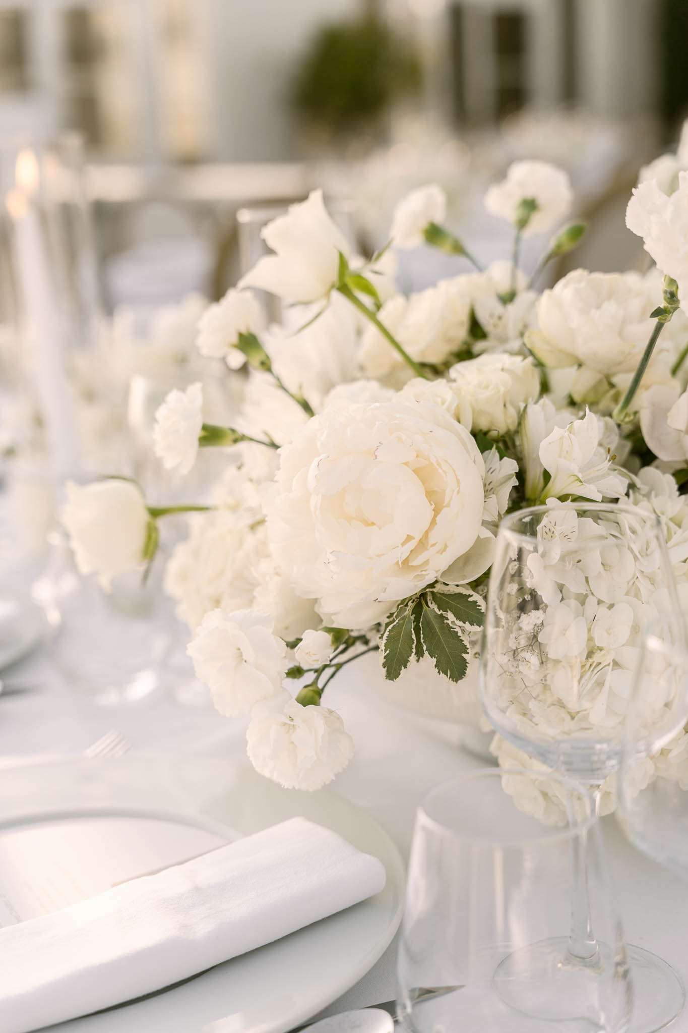 A close-up detail shot of a wedding reception table setting with an all-white floral centerpiece featuring ivory garden roses, white peonies, white hydrangeas, sweet peas, and small spray carnations with green foliage accents. The table is dressed in a white linen tablecloth with white porcelain plates, a folded white linen napkin, silver cutlery, and clear crystal glassware. In the background, tall white taper candles in slim glass holders and additional table settings are softly blurred, indicating a larger reception setup. The overall decor palette is a strict white-on-white scheme with touches of green, consistent with a modern classic or minimalist styling approach.
