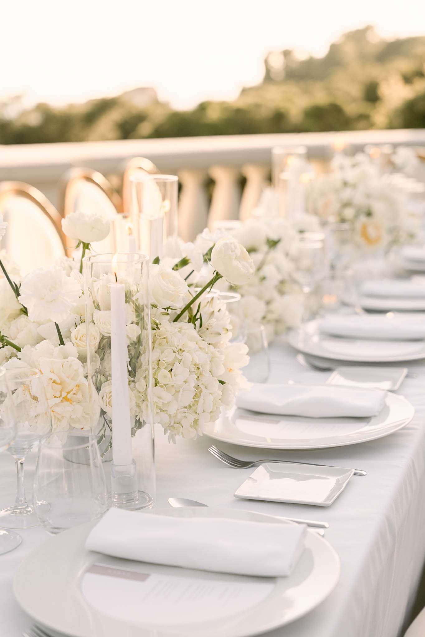Close-up detail shot of an outdoor wedding reception table set on what appears to be a terrace or balustrade-lined terrace, photographed in warm golden evening light. The table is dressed in a white linen tablecloth with all-white place settings including stacked white porcelain plates, white folded napkins, silver cutlery, and clear glassware. The centerpiece runs the length of the table and features low, lush arrangements of white hydrangeas, white garden roses, white ranunculus, and white peonies, interspersed with tall white taper candles in clear glass hurricane holders. Gold-rimmed charger plates are visible on chairs in the background, and the overall styling palette is entirely white and ivory with clear glass accents, creating a monochromatic, minimalist-modern aesthetic. The long table recedes into soft background blur, emphasizing the repeating place settings and floral runners.
