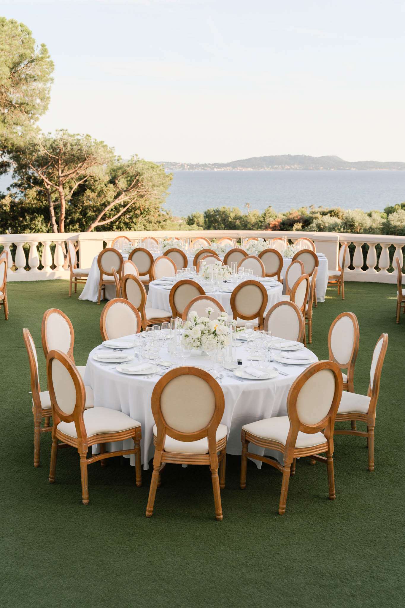 An outdoor wedding reception setup on a manicured lawn terrace overlooking the sea, with a white balustrade railing bordering the perimeter. Three round tables are visible, dressed in white floor-length tablecloths and set with white plates, folded napkins, and multiple wine glasses per place setting. Each table features a low centerpiece of white flowers — appearing to be white roses and similar blooms — arranged in a compact cluster. The chairs are Louis XVI-style with natural wood frames and cream upholstered seats and oval backs, with approximately ten chairs per round table. The decor palette is entirely white and natural wood, giving a classic, clean aesthetic. The shot is a wide-angle portrait-orientation image taken from a slightly elevated angle, capturing the full table setup in the foreground with the sea view and distant hills visible beyond the balustrade. Potential venue feature image.
