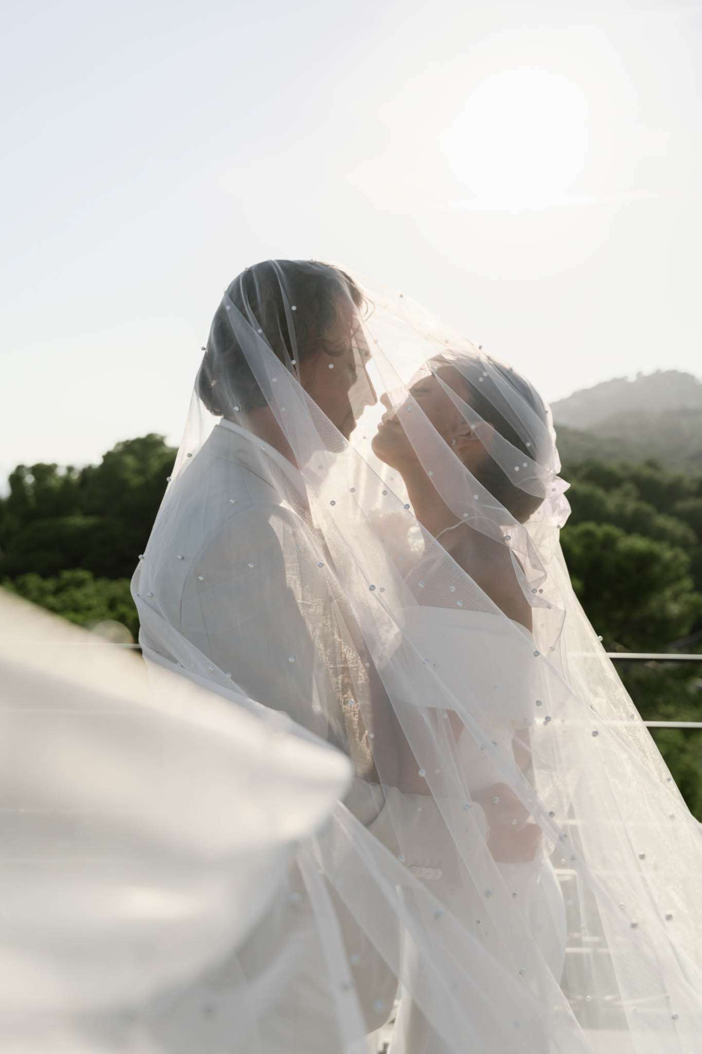 Couple shares near-kiss portrait draped under pearl-embellished cathedral veil billowing in backlit natural light