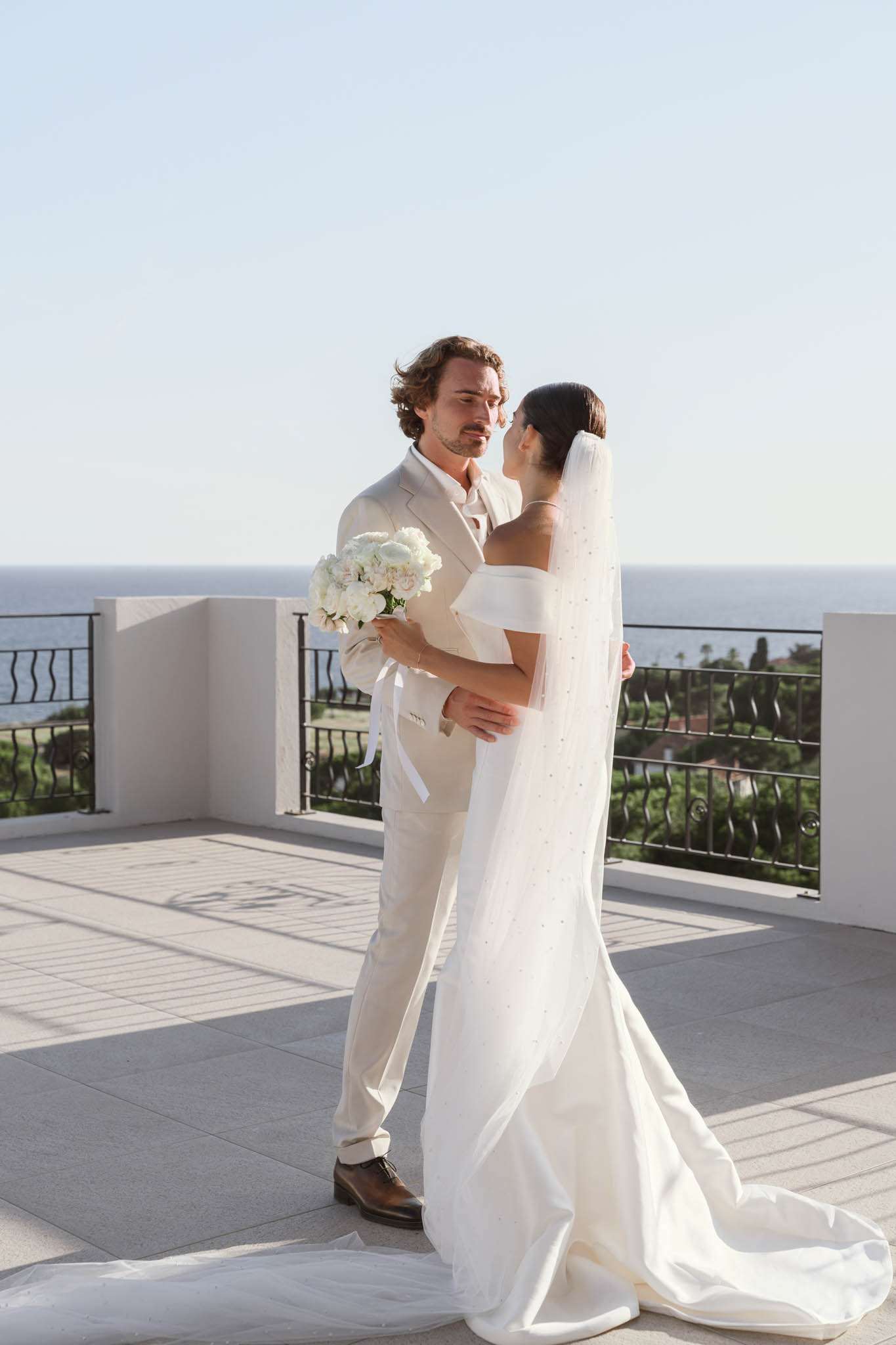 Bride with pearl-embellished veil and peony bouquet embracing groom in cream suit on sea-view terrace