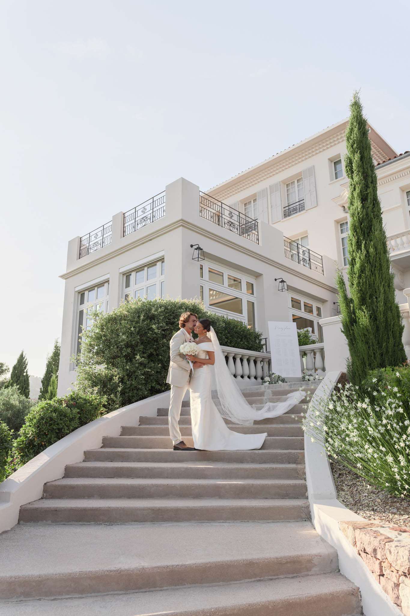 Couple on grand staircase of white villa with cathedral veil trailing down steps