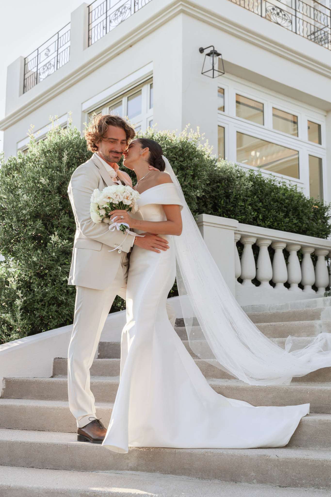 A couple portrait shot outdoors on the stone staircase of a white classical-style villa or manor, featuring wrought-iron balconies, large windows, and a white balustrade with turned columns. The bride wears a fitted off-the-shoulder white satin gown with a long cathedral-length veil and holds a round bouquet of white peonies and white roses with a white ribbon tie. The groom wears a light sand/beige suit with a blush-pink tie and brown leather dress shoes. The two are embracing and nearly kissing, with the bride leaning into the groom. The overall styling is clean and modern with an all-white and ivory palette. Full-length portrait composition shot in natural daylight. Potential venue feature image.