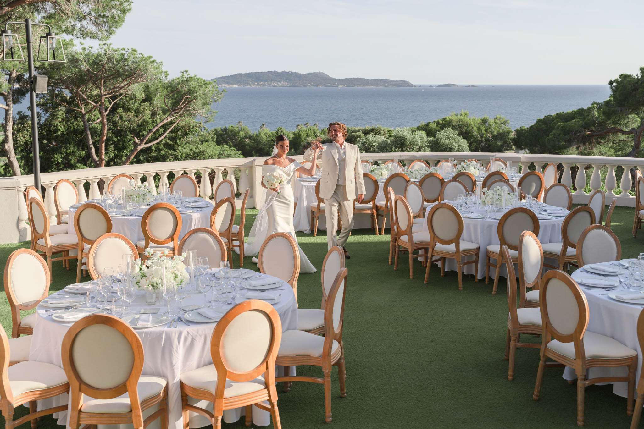 A bride and groom walk through a fully set outdoor reception terrace, pausing to take in the space before guests arrive. The bride wears a fitted off-the-shoulder ivory gown with a train and holds a white bouquet, while the groom wears a light beige suit. The terrace features round tables dressed in white linen, set with glassware, white plates, and low centerpieces of white blooms — likely white roses and green foliage. Seating consists of natural wood Louis XVI-style chairs with cream upholstered backs and seats. The terrace is bordered by a white balustrade railing and overlooks a panoramic sea view with a pine-covered island in the distance. The overall décor palette is an all-white and natural wood scheme with a classic, clean aesthetic. Wide shot capturing the full table layout and couple in the middle ground. Potential venue feature image.