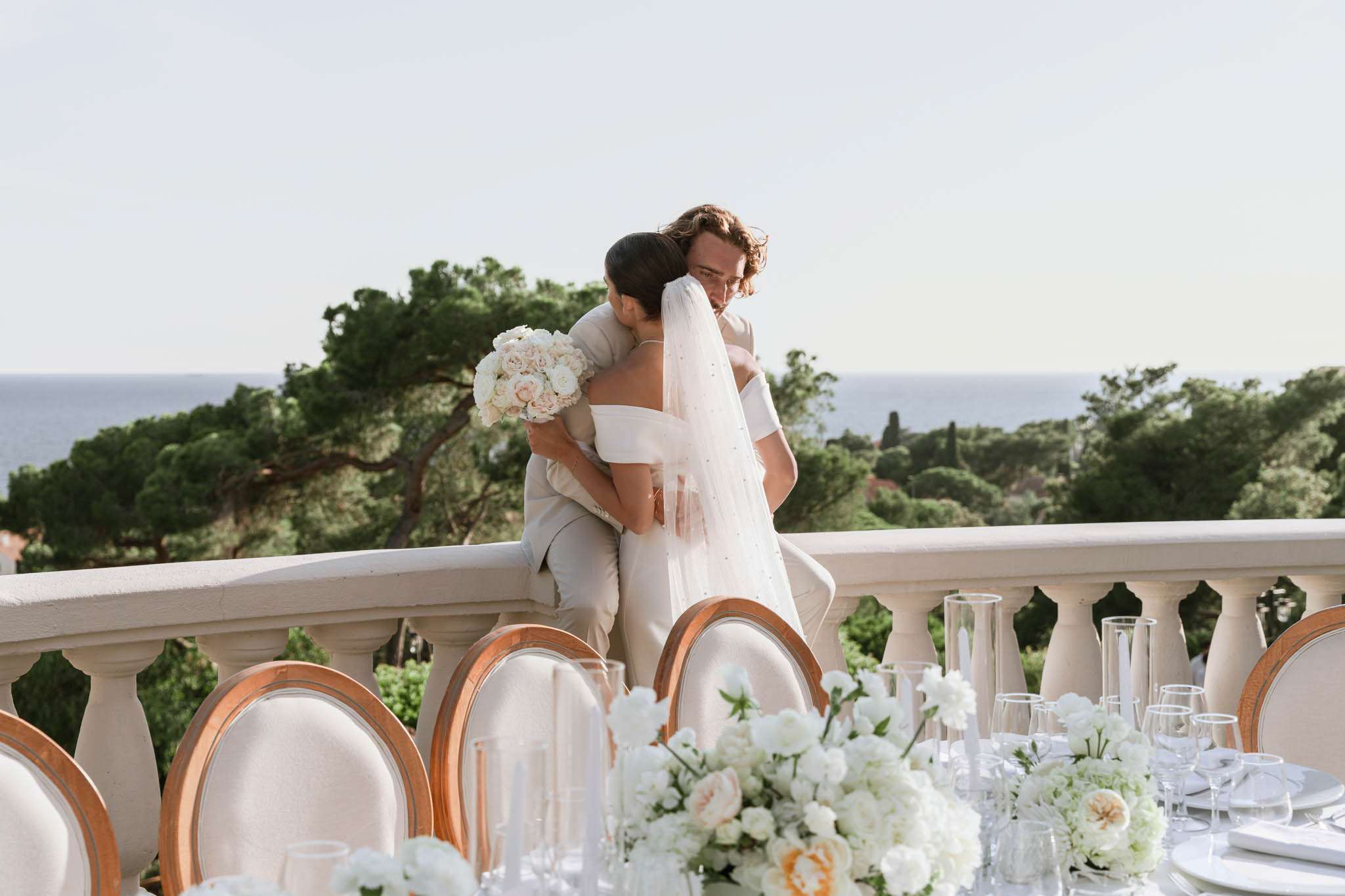 Couple embracing on sea-view terrace with styled reception table of white ranunculus in foreground