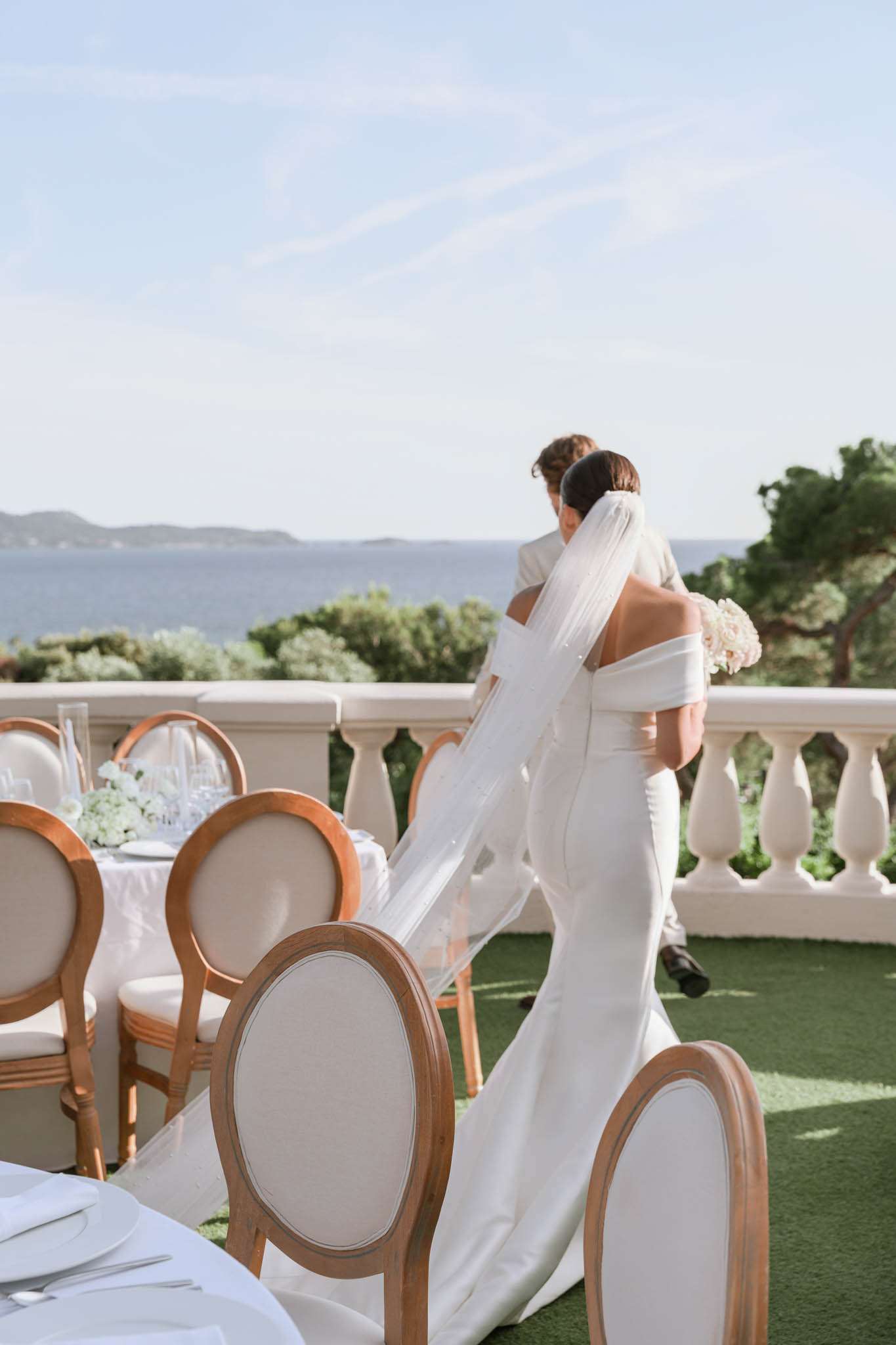 A bride and groom embrace on an outdoor terrace overlooking a sea view, captured from behind in a mid-distance portrait shot. The bride wears a fitted off-the-shoulder white gown with a long cathedral veil and holds a bouquet of blush and white blooms, likely hydrangeas and roses. The groom is dressed in a light beige or ivory suit. In the foreground, a dressed reception dining table is partially visible, set with white linens, glassware, and low white floral centerpieces, surrounded by Louis XVI-style chairs with natural wood frames and ivory upholstered oval backs. The terrace features a white balustrade railing, and the overall decor palette is white and natural wood with a clean, classic aesthetic.