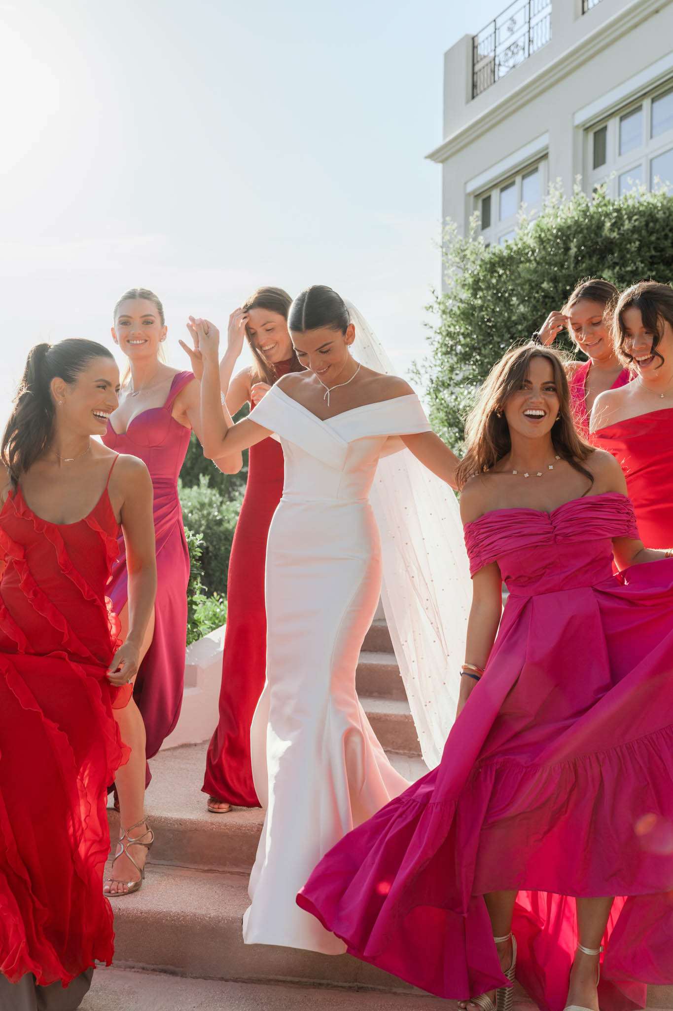 Bride in white gown surrounded by six bridesmaids in mismatched red and fuchsia dresses laughing on villa steps