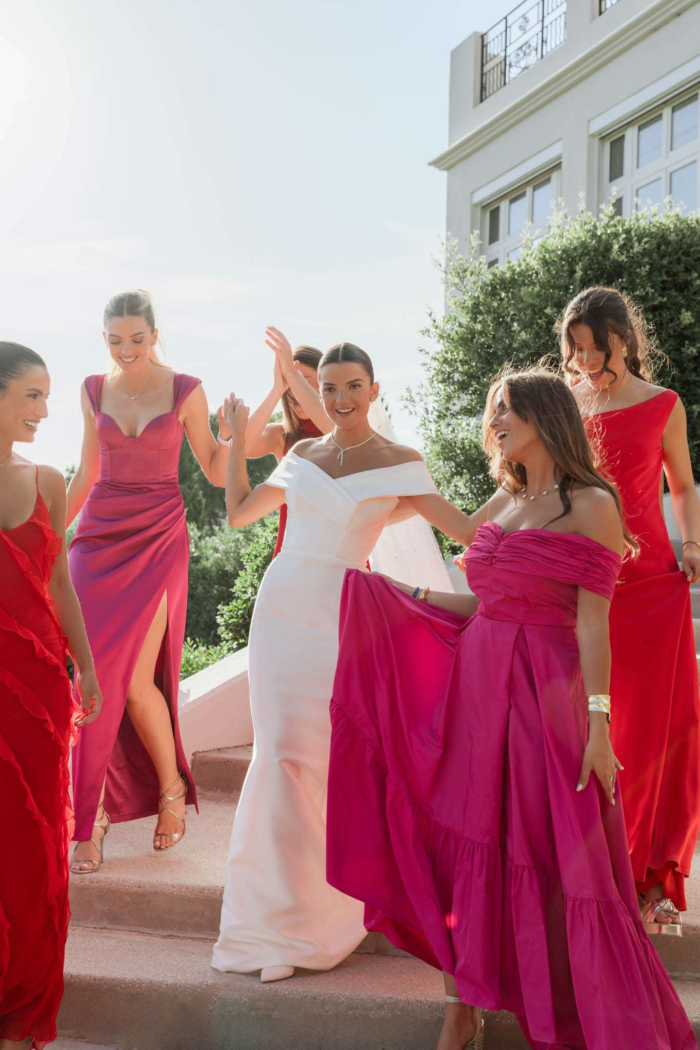 Bride laughing on stone steps with five bridesmaids in bold red and hot pink mismatched gowns
