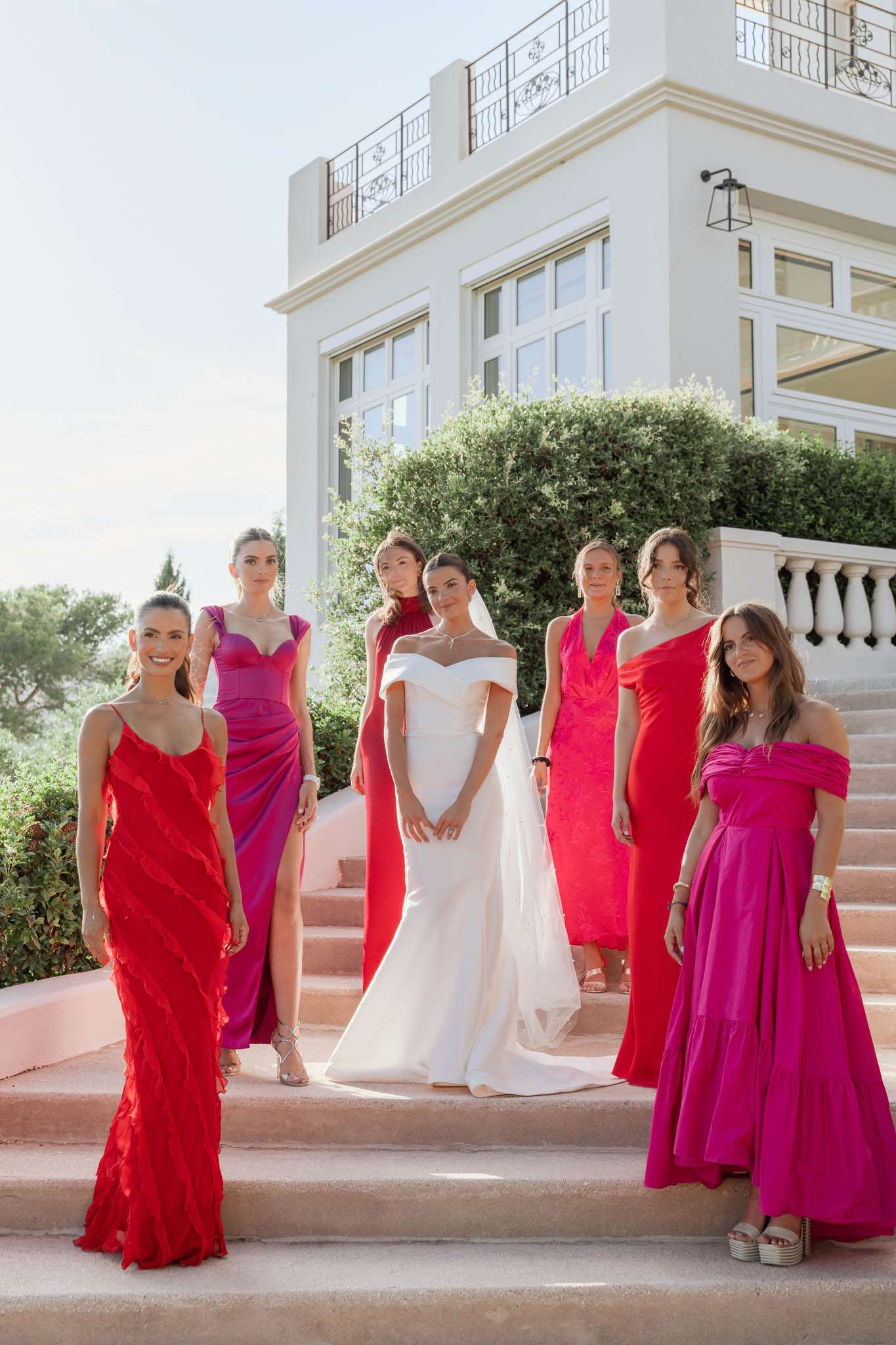 A bridal party portrait taken outdoors on the steps of a white villa with wrought-iron balconies and large windows, shot in bright natural daylight. The bride stands at the center wearing an off-the-shoulder white fitted gown with a cathedral-length veil and a diamond necklace, surrounded by six bridesmaids. The bridesmaids wear a mix-and-match color palette of red and fuchsia/magenta floor-length gowns in varying silhouettes — including a ruffled red spaghetti-strap dress, a purple-to-pink ombré draped gown with a slit, a red turtleneck halter dress, a red wrap-style dress, a red one-shoulder gown, and a bright fuchsia off-the-shoulder tiered dress. The styling theme is bold and modern with a saturated red and pink color scheme. The shot is a medium-wide portrait with the group arranged across two steps of the terrace staircase.