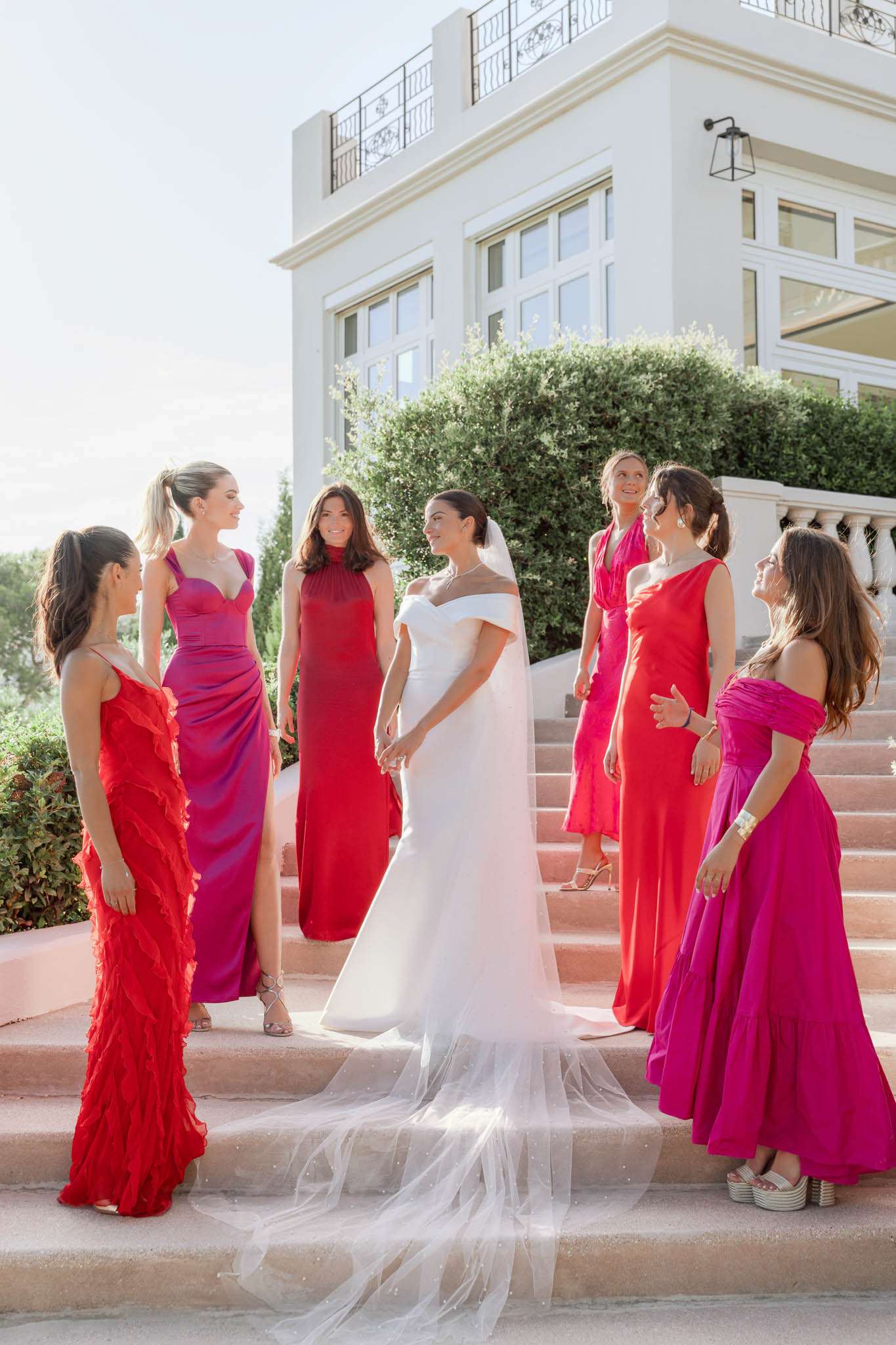 A bridal party portrait taken outdoors on the stone steps of a white render villa with wrought-iron balcony railings. The bride stands at the center wearing a fitted off-the-shoulder white gown with a long cathedral-length tulle veil that cascades down the steps. She is surrounded by six bridesmaids in mismatched floor-length gowns across a red, fuchsia, and magenta color palette — styles include a red ruffled spaghetti-strap dress, a purple-to-magenta satin ruched gown, a high-neck deep red dress, a coral-red halter dress, a bright fuchsia off-the-shoulder dress, and a hot pink one-shoulder gown. The group is arranged informally on the steps, facing inward toward the bride and interacting with one another in a candid, conversational pose. The overall styling theme is bold and modern with a vivid warm color palette. Wide mid-shot composition.