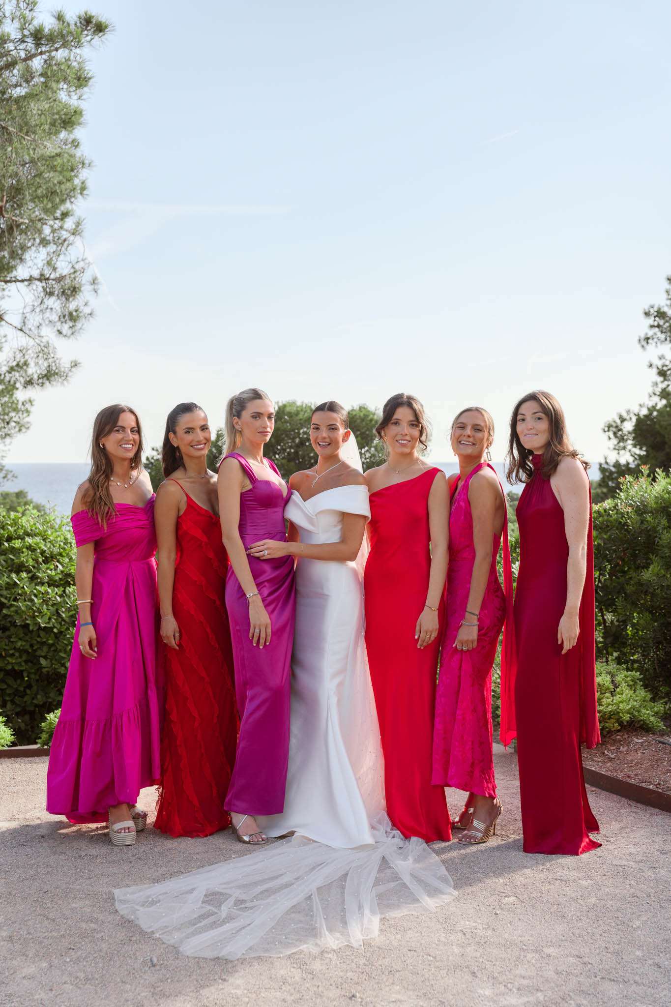 A bridal party portrait taken outdoors on a gravel terrace with a sea view visible in the background. The bride stands at center wearing a white off-the-shoulder fitted gown with a long tulle veil trailing on the ground. She is flanked by six bridesmaids wearing mismatched floor-length gowns in a coordinated palette of fuchsia, magenta, red, and deep crimson — styles include an off-the-shoulder tiered fuchsia dress, a red slip dress, a purple-magenta satin gown, a one-shoulder red dress, a hot pink halter dress, and a high-neck dark red gown. The group poses in a loose line facing the camera, smiling. The setting appears to be a Mediterranean coastal venue, suggested by the sea horizon visible between the trees in the background. This is a wide group portrait shot in natural daylight.
