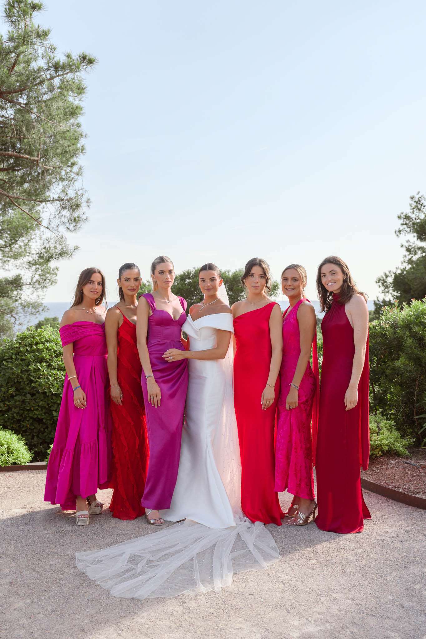 Bride in white gown with cathedral veil flanked by six bridesmaids in red and pink satin dresses on a seaside terrace