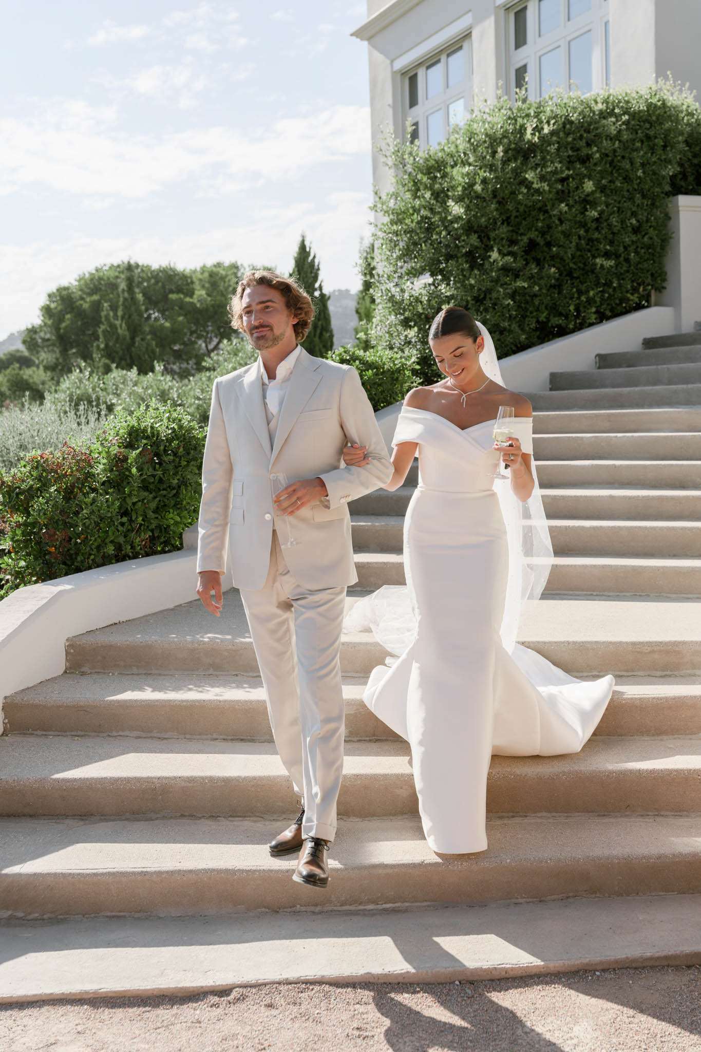 A couple portrait taken outdoors on the wide stone staircase of a white-rendered villa or château, shot in natural daylight. The bride wears a fitted off-the-shoulder white gown with a cathedral-length train and a long sheer veil, accessorized with a delicate necklace; she holds a champagne flute and smiles downward as they descend. The groom wears a light sand-beige suit with a white shirt and no tie, paired with brown Oxford shoes. The composition is a full-length portrait with the venue's white-rendered façade and manicured hedging visible in the background, reflecting a clean, modern classic styling aesthetic.