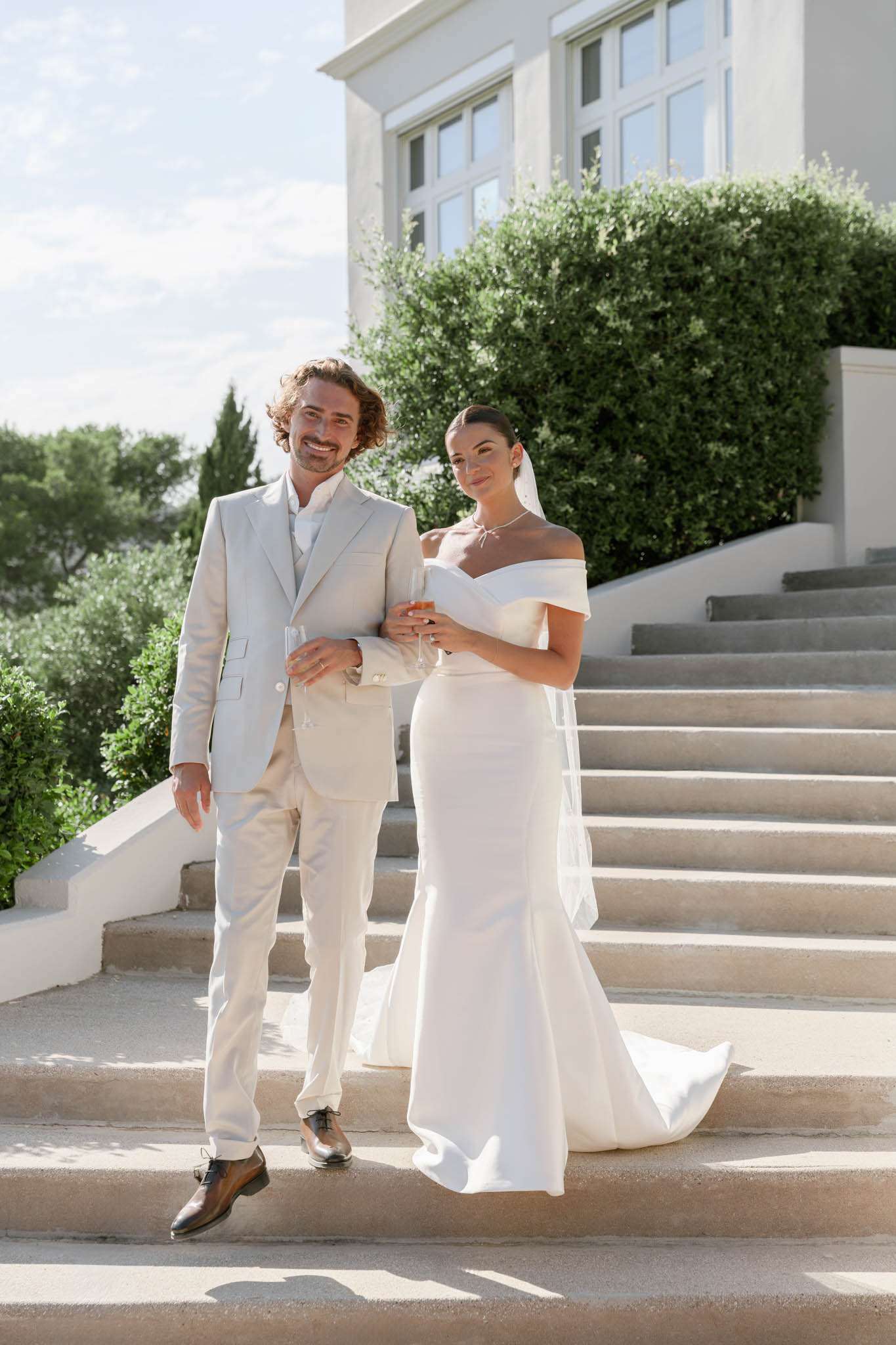A couple poses together on the stone steps of a white-rendered villa or château during what appears to be a cocktail hour or portrait session. The groom wears a light sand-beige three-piece suit with brown leather Oxford shoes and holds a champagne flute, while the bride wears a fitted off-the-shoulder ivory crepe gown with a chapel train and a long sheer veil, and holds a glass of rosé. The styling is clean and modern with a minimal, monochromatic palette across both outfits. The shot is a full-length portrait taken in bright natural daylight with the venue's white architecture and neatly trimmed hedging visible in the background.