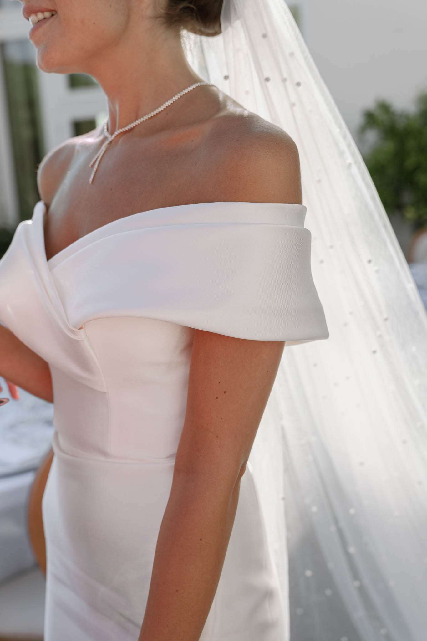 Close-up of bride in off-the-shoulder satin gown with pearl necklace and crystal-dotted veil