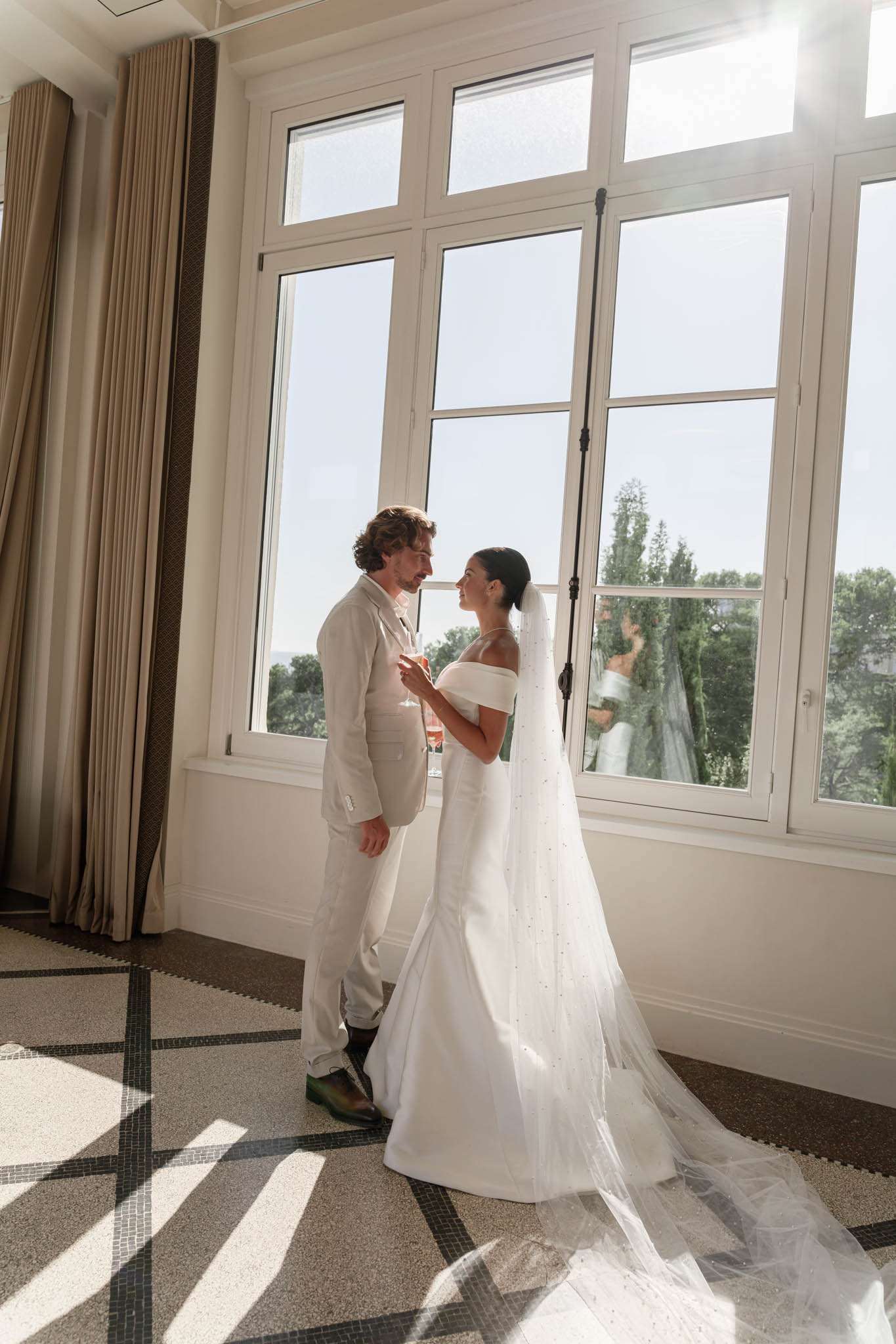 A couple portrait taken indoors in a bright room with floor-to-ceiling white-framed windows that flood the space with natural sunlight. The bride wears an off-the-shoulder white fitted gown with a long cathedral-length veil scattered with small crystal or pearl embellishments trailing across the patterned mosaic tile floor. The groom is dressed in a light beige suit with no tie and an open collar, and holds what appears to be a glass of rosé. The two face each other closely, smiling, in a candid and relaxed pose. The room features a classic interior with cream drapery, white molding, and a geometric mosaic tile floor in neutral and dark tones, suggesting an upscale hotel or château setting. The composition is a full-length portrait shot with strong backlight creating a warm, sun-drenched atmosphere.