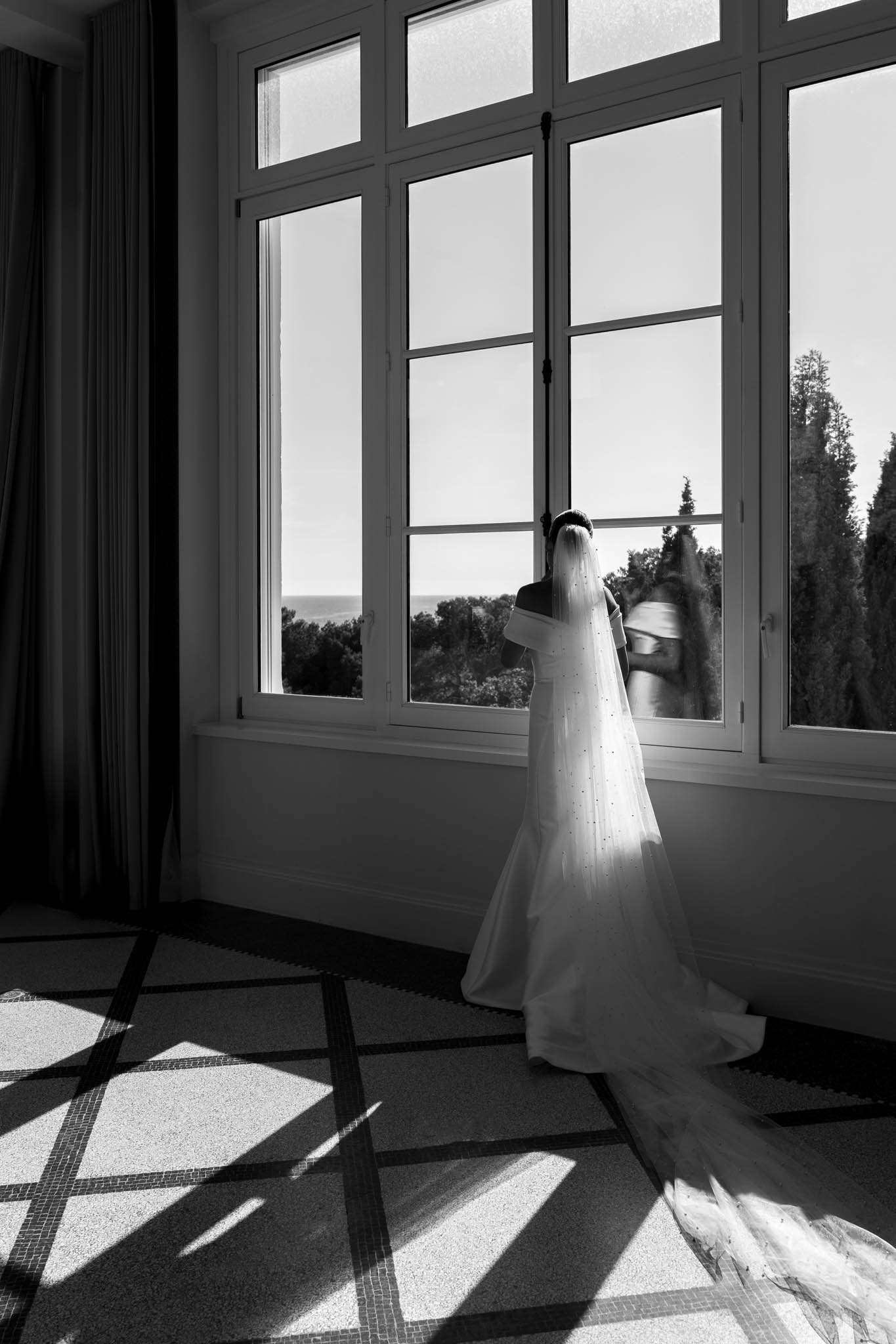 Black-and-white portrait of bride in off-shoulder gown and cathedral veil looking through French windows