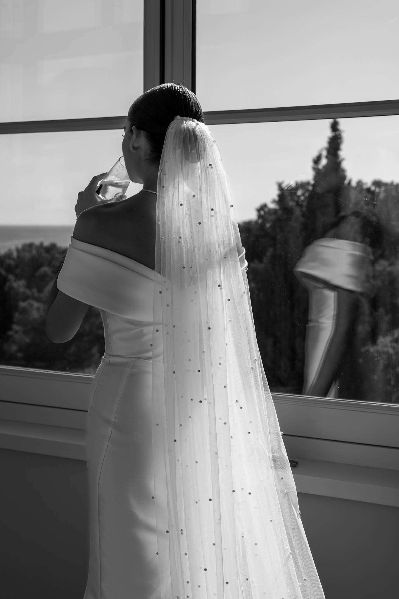 Bride sips champagne at floor-to-ceiling window with pearl-scattered cathedral veil from behind in B&W