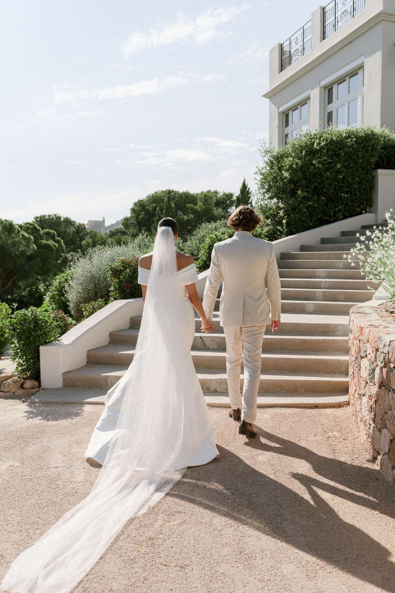 A couple portrait shot from behind showing the bride and groom walking hand-in-hand toward a wide stone staircase leading up to a white rendered villa or manor house. The bride wears an off-the-shoulder white fitted gown with a long cathedral-length veil that extends to the ground, with subtle embellishment visible on the veil fabric; her dark hair is worn up. The groom is dressed in a light sand/cream suit with brown dress shoes. The setting is outdoor on a gravel terrace with manicured hedges and Mediterranean-style landscaping surrounding the staircase. The shot is a full-length portrait taken in natural daytime light, with the architectural facade of the venue prominently featured in the upper right of the frame. Potential venue feature image.