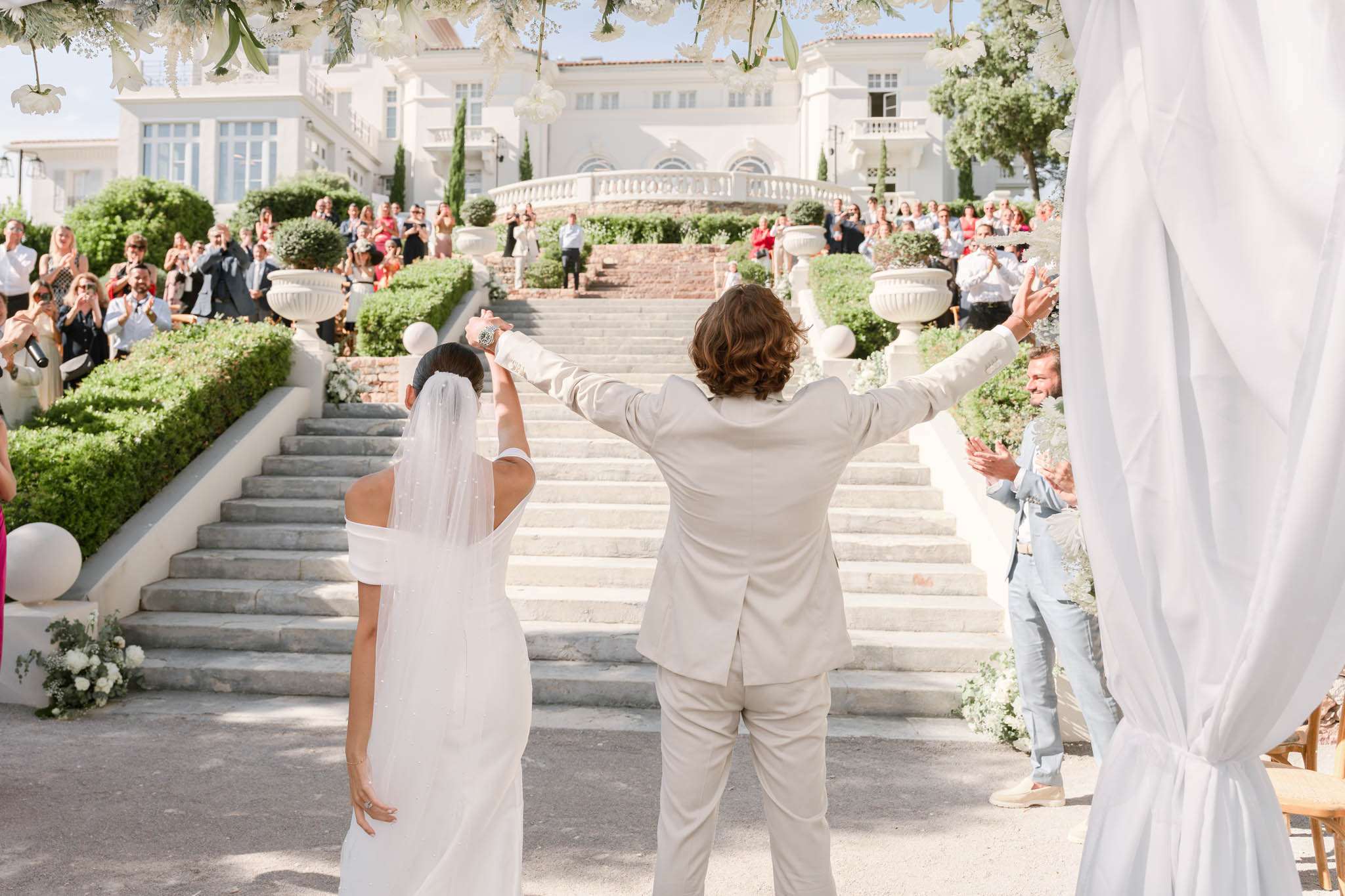 Couple raises joined hands as 70 guests applaud on tiered stone stairs of white villa with floral arch